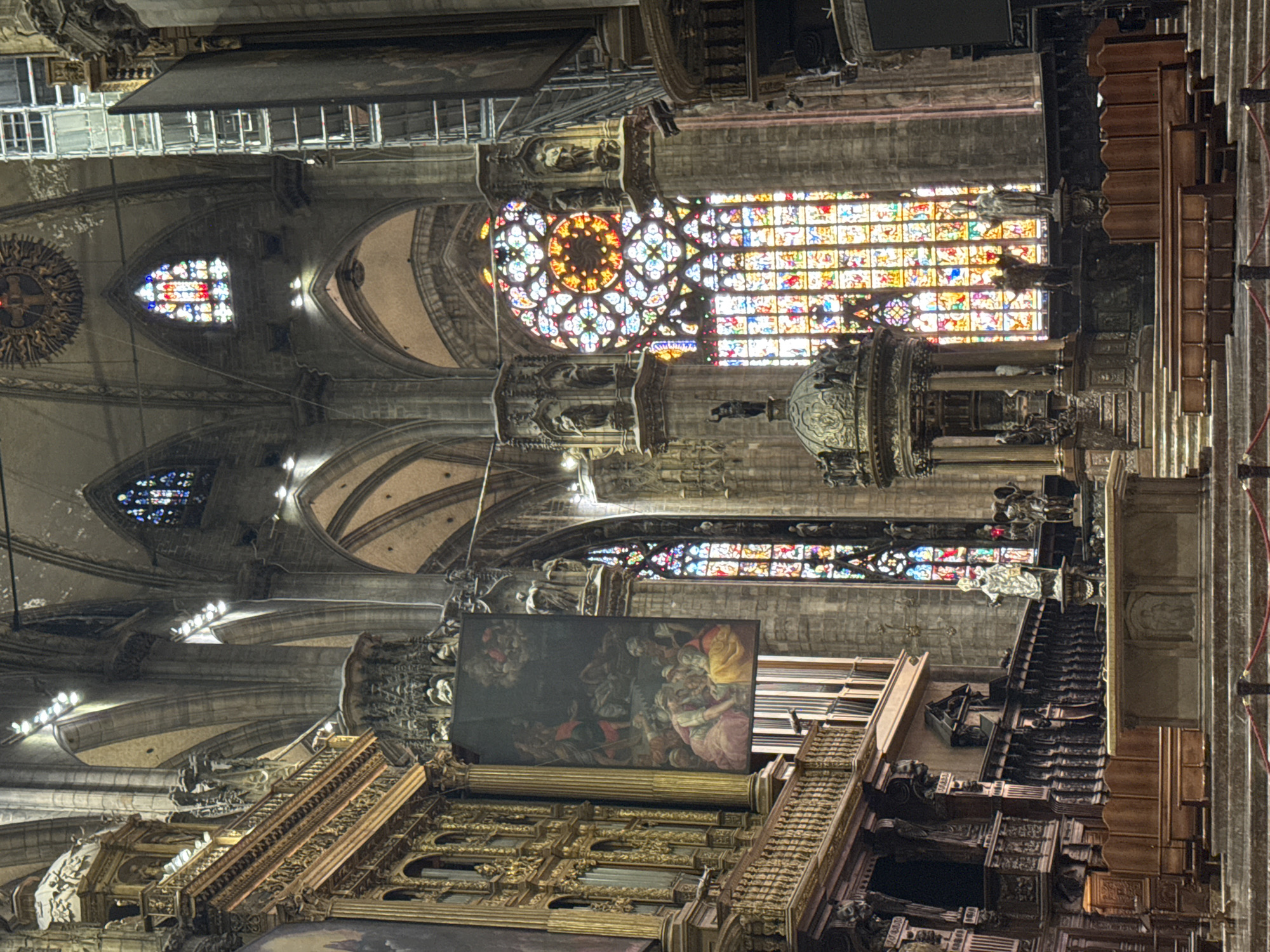 The image shows the interior of the Milan Cathedral, also known as the Duomo di Milano, located in Milan, Italy. The scene depicts the nave of the cathedral, with its soaring Gothic architecture. Dominating the view are large, intricate stained-glass windows, casting colorful light into the space. In the foreground, there are rows of ornate wooden pews and a large altar area. To the left, a grand organ is visible, and a large painting hangs suspended. Statues and decorative architectural elements are present throughout the scene, highlighting the rich history and artistry of the cathedral. The overall impression is one of grandeur and solemnity, with natural light filtering through the vibrant glass.