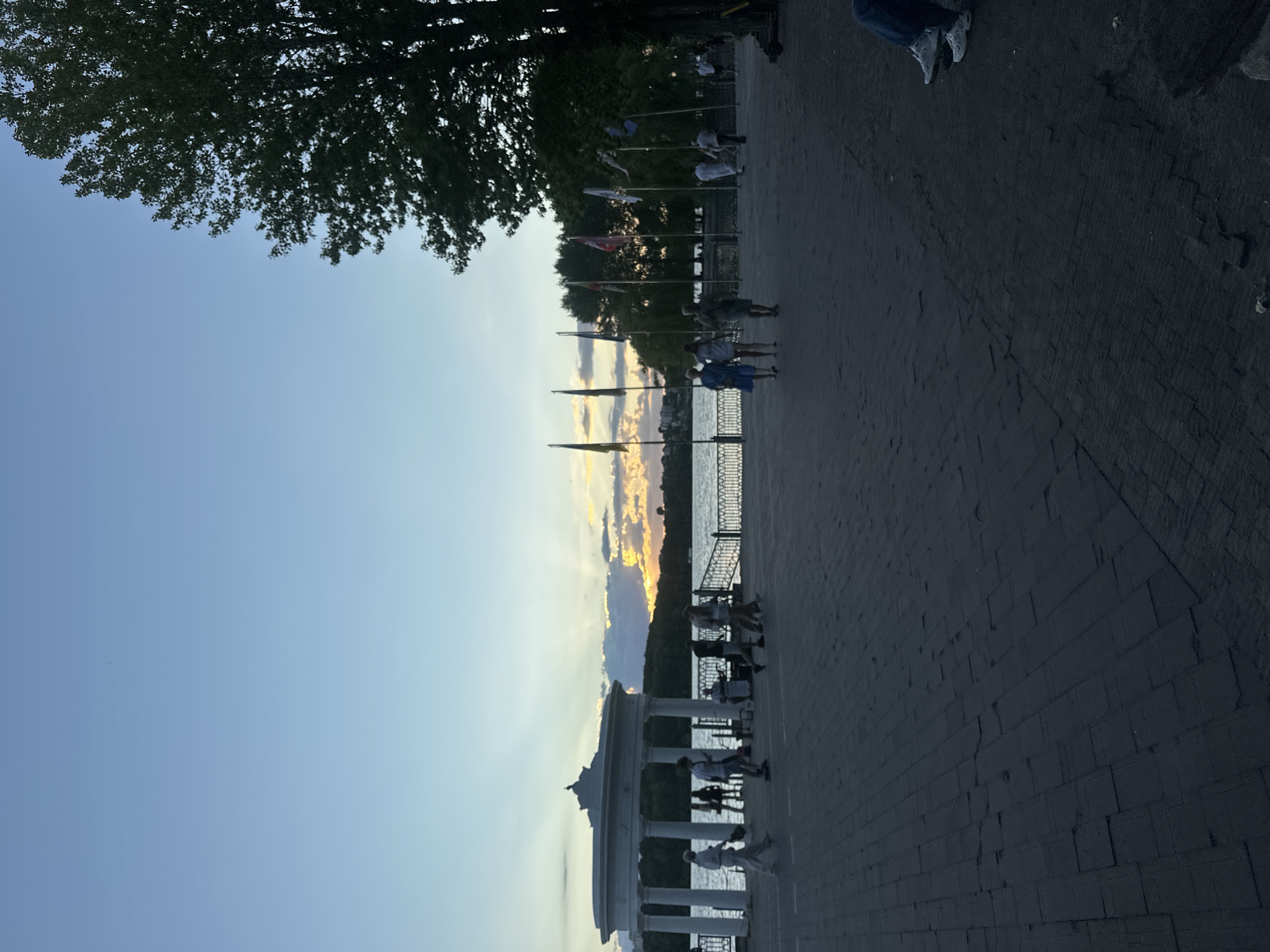 The image depicts an outdoor scene at dusk, likely on a waterfront promenade in Ternopil, Ukraine. The sky displays a gradient of blue, transitioning to warmer tones of orange and yellow near the horizon where the sun is setting. Clouds are present, with some illuminated by the setting sun, casting rays of light.
In the mid-ground, a body of water, possibly Ternopil Lake, is visible. Along the water's edge, a white gazebo-like structure with columns stands prominently on the left. A railing separates the promenade from the water. Several flags on poles are planted in a line further to the right, behind a low fence. Trees with dense foliage are seen in the background, some silhouetted against the sky.
On the paved promenade, several people are walking and standing. On the far left, a group of individuals are seen near the gazebo, some appearing to be walking. Further to the right, a few people are walking along the promenade, including a woman in a blue dress and another in a white top and skirt. To the right of the flags, more people are visible, some in the distance. In the foreground, the promenade is paved with dark rectangular stones arranged in a pattern. On the far right, a person's legs and feet, clad in blue jeans and white sneakers, are visible as they sit on the edge of the frame. The overall atmosphere suggests a public gathering place during an evening stroll.