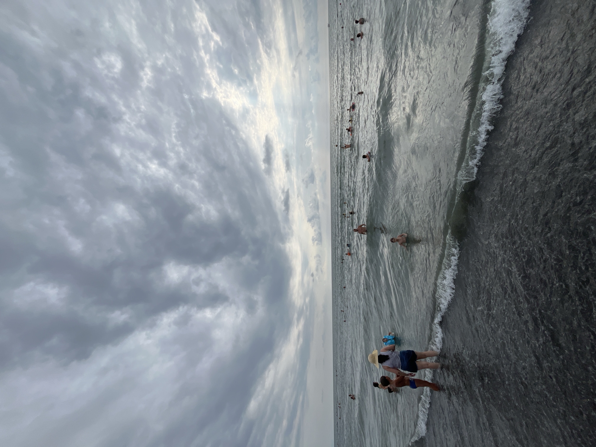 The image depicts a beach scene in Ureki, Georgia, under a cloudy sky. Numerous people are visible in the water, with some wading near the shore and others further out. Among the figures in the foreground, a woman wearing a straw hat and a striped top stands with her back to the camera, facing the sea. A young boy in blue swim trunks stands beside her. Other individuals are scattered throughout the water, engaged in swimming and wading. The ocean is calm with small waves breaking near the shore. The dark, textured sand of the beach is visible in the immediate foreground. The overall lighting suggests it may be overcast or late in the day.
