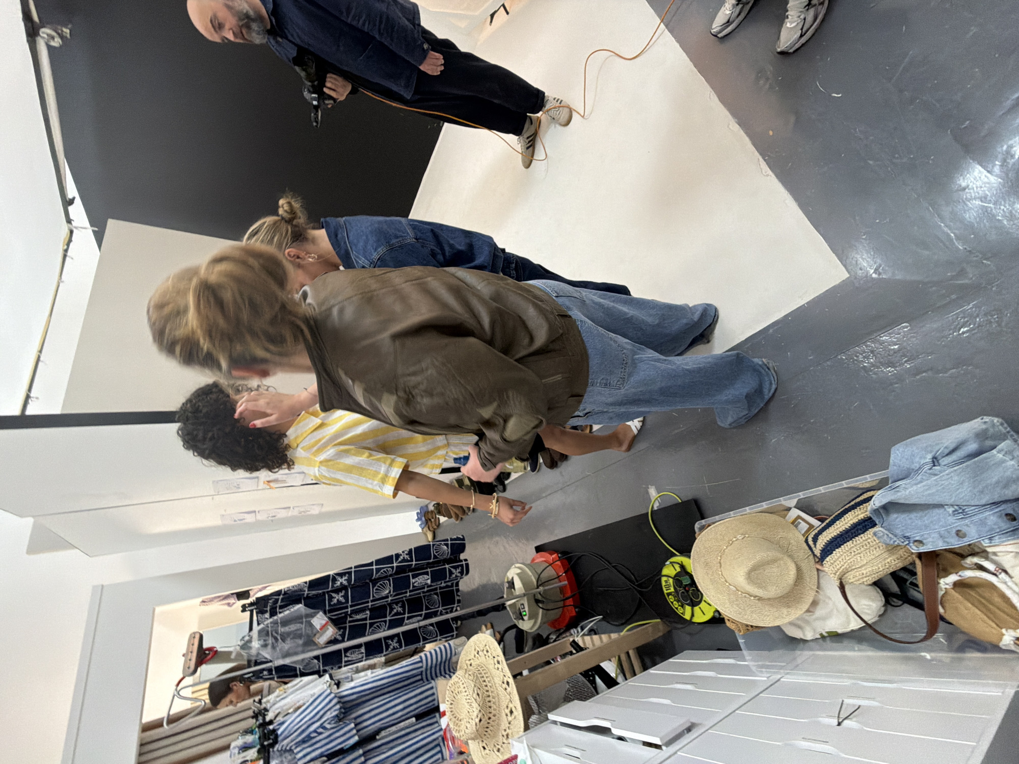 The image shows the interior of a photography studio. A photographer is capturing images of a scene with three individuals. One man is photographing the other two, who appear to be models. The models stand on a white and black backdrop. The person taking the photograph is standing at an angle to the models with his arms raised and holding a camera to his eye. There is equipment on the right, including a power strip and what looks like a red heater. The location and existence of the city of Manchester are not shown in the image.