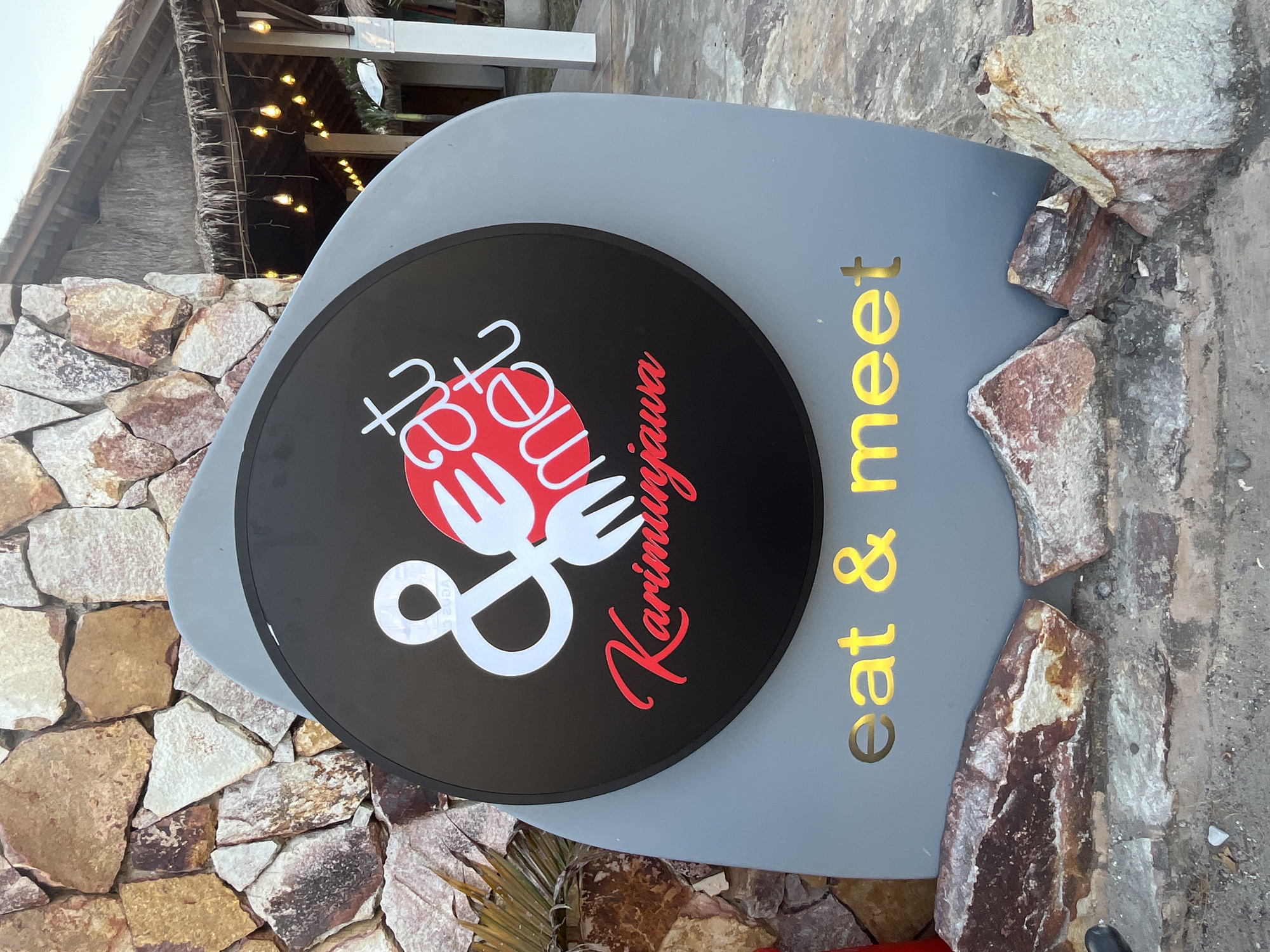 The image shows a sign for "Eat & Meet Karimunjawa" located in Karimunjawa, Indonesia. The sign is circular and black with white and red lettering, featuring a stylized ampersand and forks. Below the circular sign, a gray, curved panel displays the words "eat & meet" in illuminated yellow lettering. The sign is partially surrounded by rocks and is positioned in front of a building with a thatched roof and string lights. The background shows a stone wall.