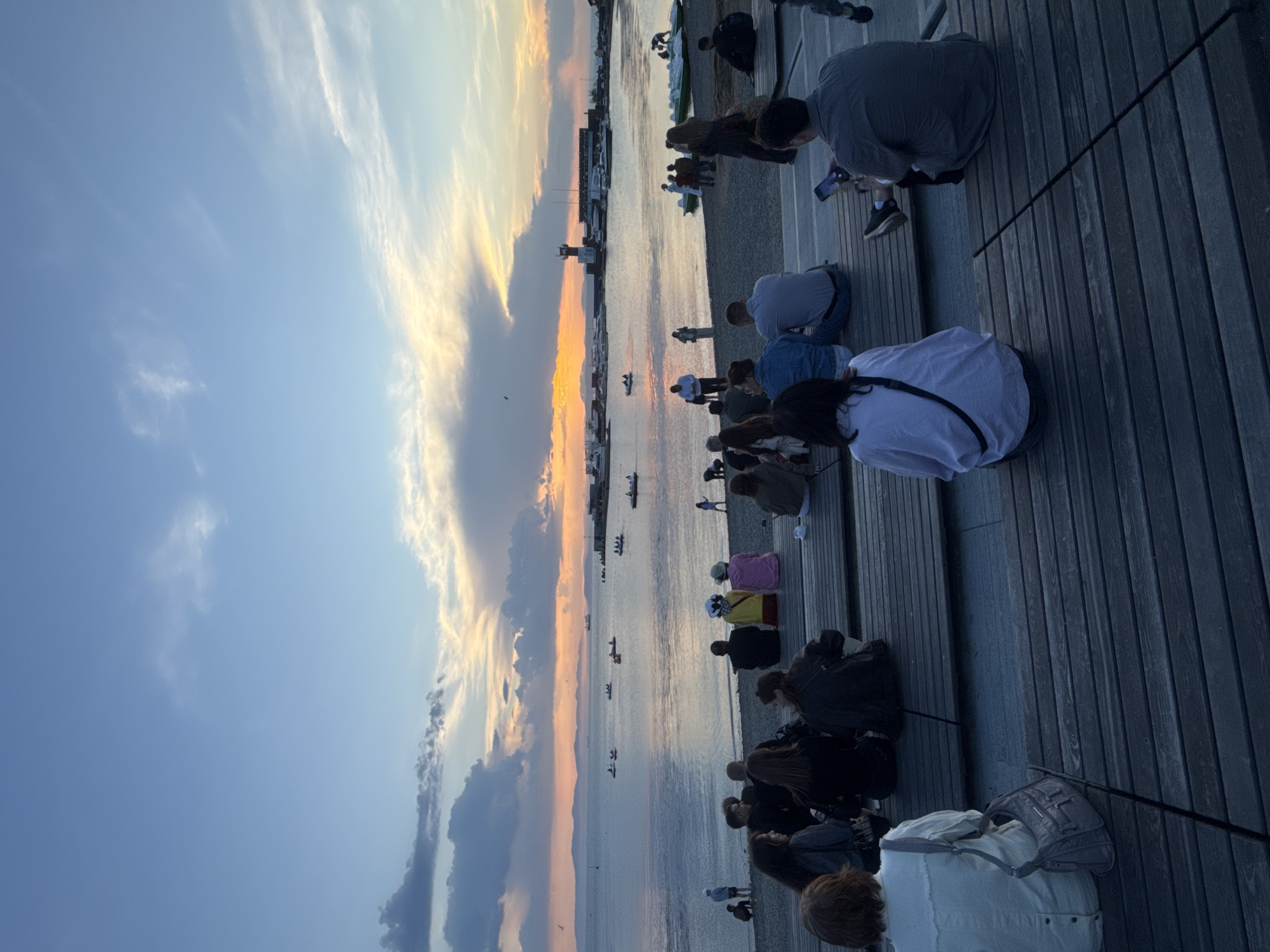 The image captures a view of the waterfront in Vladivostok, Russia, during sunset. A group of people are seated on wooden bleachers overlooking the sea. In the distance, several boats are visible on the water, and a lighthouse stands on the horizon. The sky is painted with hues of orange, yellow, and blue, with clouds adding texture to the scene. The overall atmosphere suggests a relaxed evening gathering by the sea.