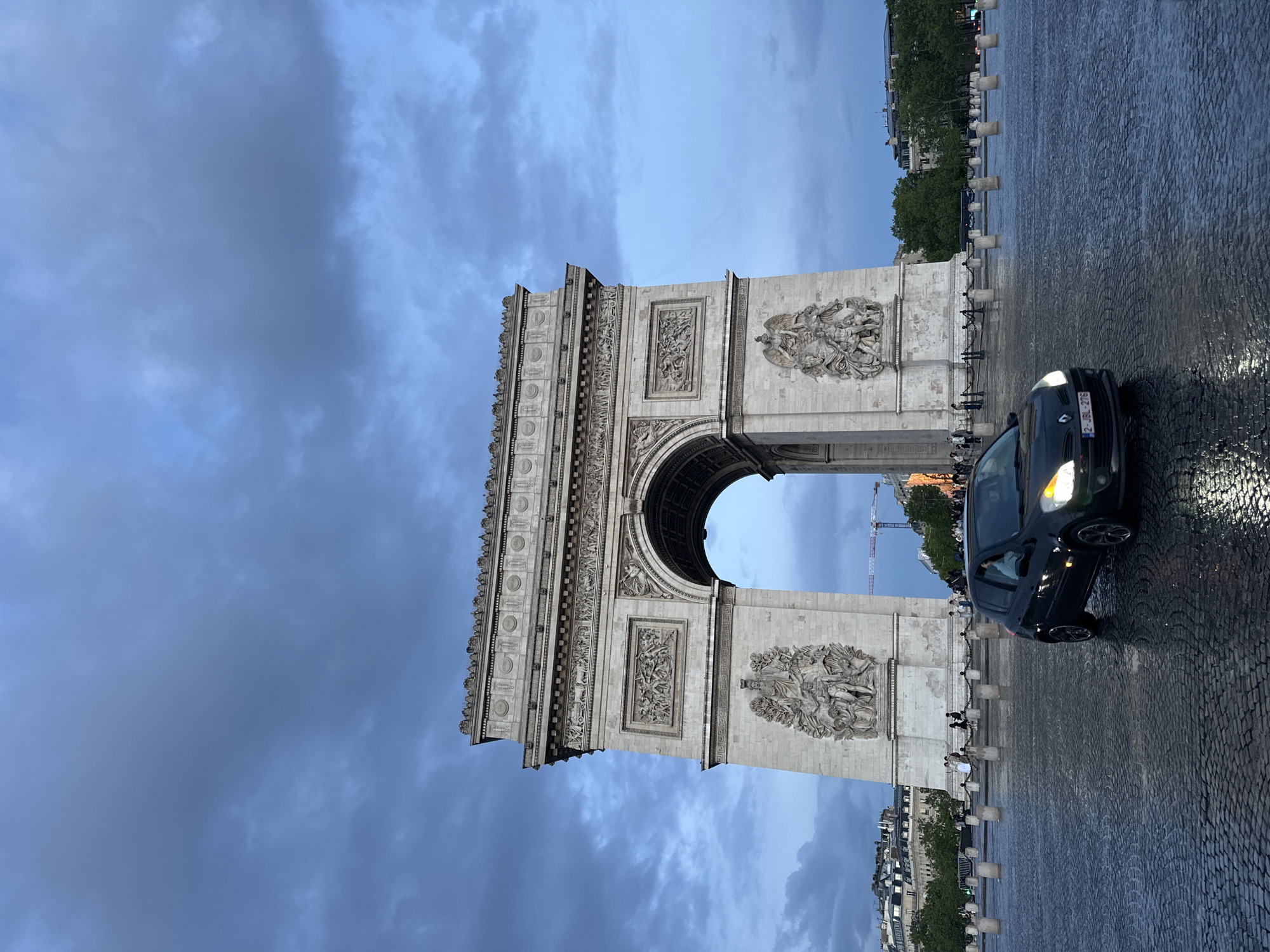 The image shows the Arc de Triomphe in Paris, France, under a cloudy sky. The ground appears wet, suggesting recent rain. A dark-colored car is prominently parked in front of the Arc, its headlights on. People are visible near the base of the Arc, but they are not the main focus. The surrounding cityscape with buildings and trees can be seen in the background.