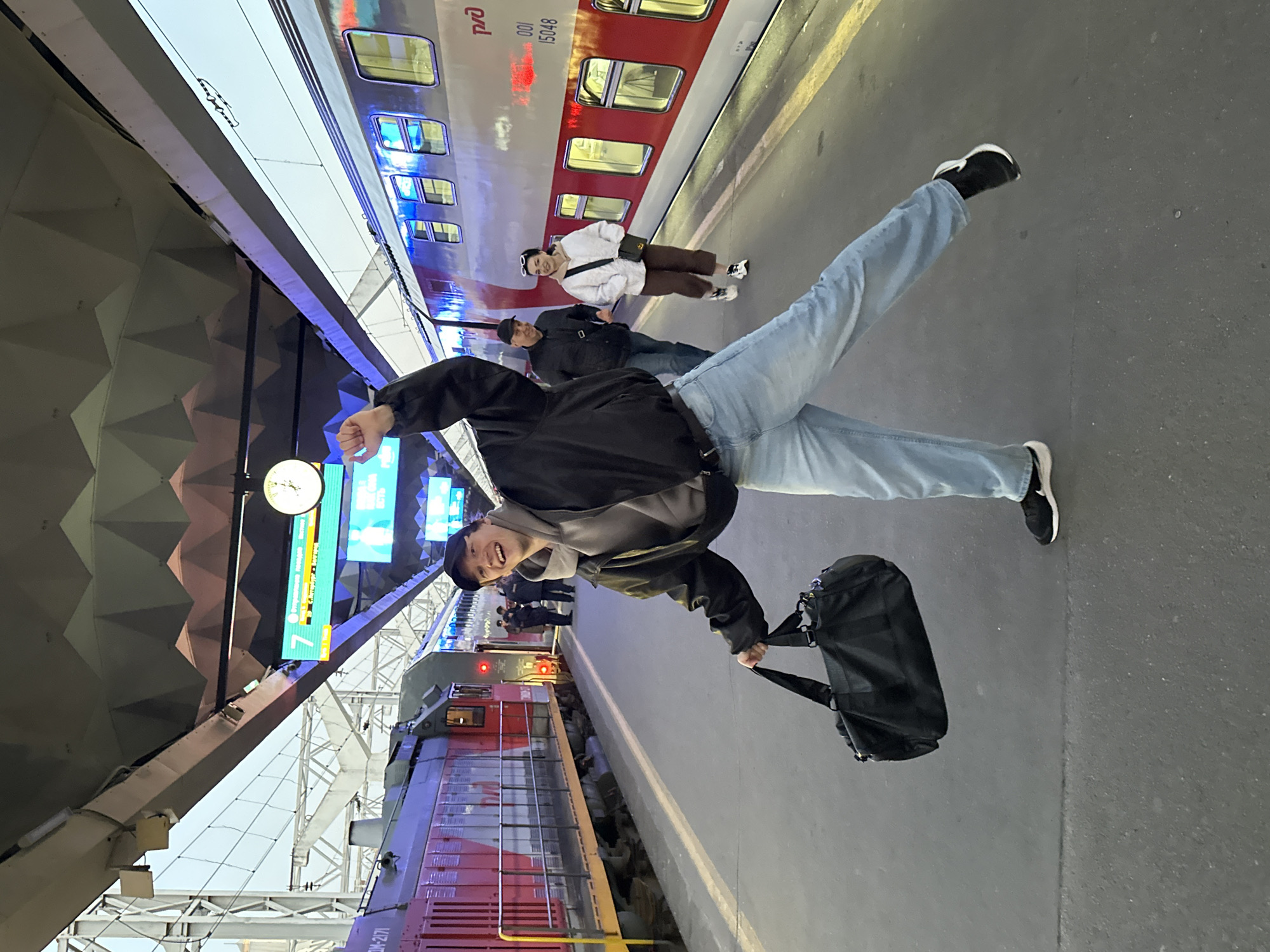A male is in the foreground striking a ballet pose on a train platform. He is wearing light blue jeans, a gray sweatshirt, and a black jacket, black shoes with white soles, and a black cap. He is holding a black bag in his right hand. His body is twisted with one leg extended towards the sky, and his other leg extended towards the ground. Other people are standing on the platform behind him. A red and white passenger train is behind the people. A red locomotive engine is behind the male subject. The train platform has a modern, angular roof structure. A sign displaying the number "7" is on the structure above. The scene is likely in Saint Petersburg, Russia, based on the design of the train and the style of the architecture.