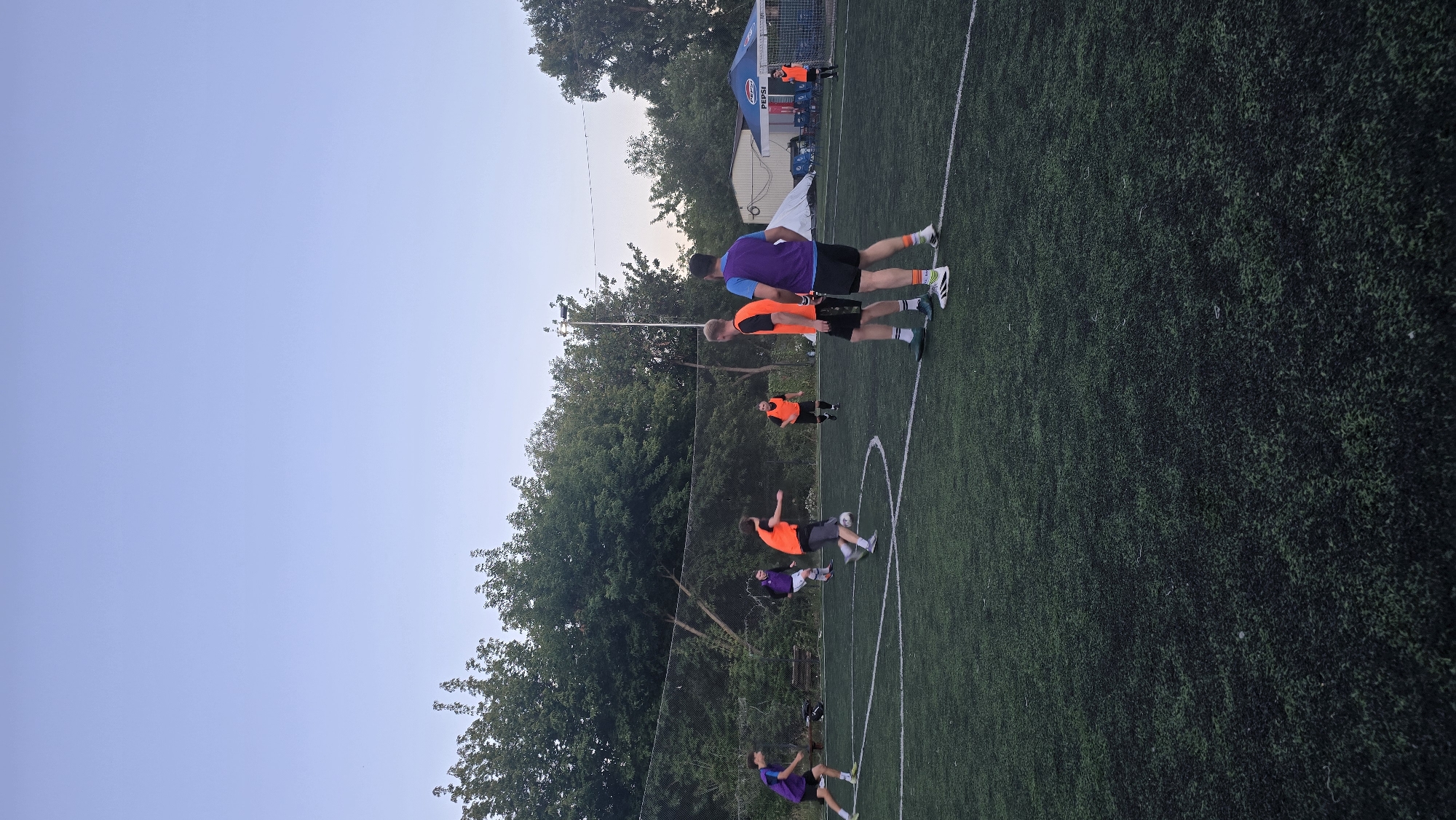 The image depicts a group of men playing soccer on an artificial turf field at dusk. The field is marked with white lines, and a soccer ball is visible near the center of the frame. Several players are wearing orange bibs, while others are in purple or blue tops and dark shorts. Some players are actively engaged in the game, with one in motion kicking the ball, and others standing and observing. In the background, trees and a fence line the edge of the field. A small white building and a blue Pepsi branded tent are also visible. The sky is a pale, clear blue, indicating early evening. The overall scene suggests a casual sports gathering. The location is identified as a sports field in Kyiv, Ukraine.