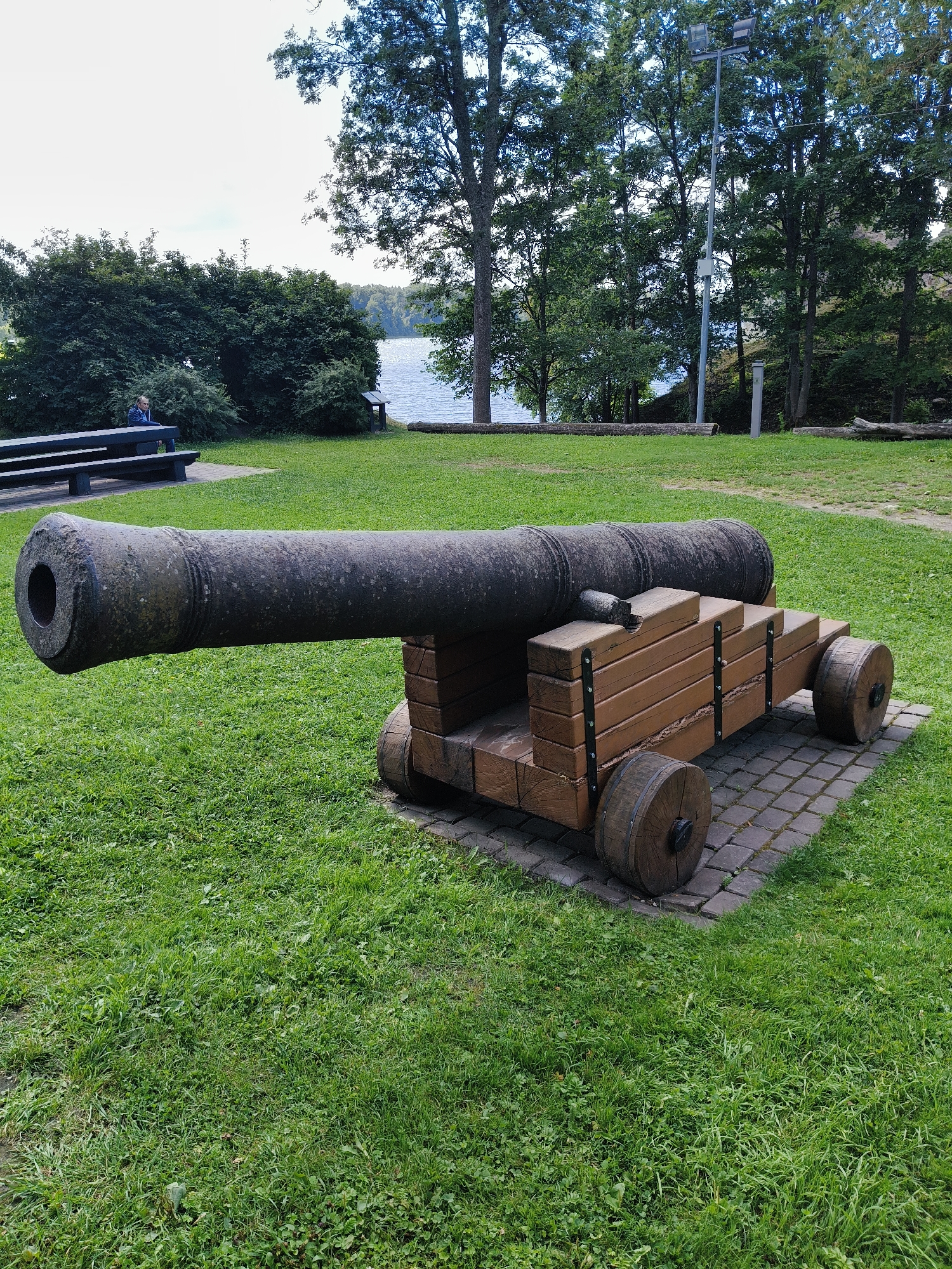 In Koknese, Latvia, a weathered, dark cannon rests on a wooden carriage with two large wooden wheels on each side. The carriage is constructed from light-colored wooden planks, stacked and reinforced with dark metal bands. The cannon and its carriage are positioned on a paved area made of gray interlocking bricks, surrounded by a lush green lawn. In the background, a body of water, likely a lake or river, is visible, bordered by trees and vegetation. A picnic table with benches is situated in the left portion of the frame, and a person is seated on one of the benches, looking away from the camera. Tall trees, some bare and some with green leaves, dominate the background, with a tall pole supporting lights and possibly security cameras standing among them. The sky is overcast.