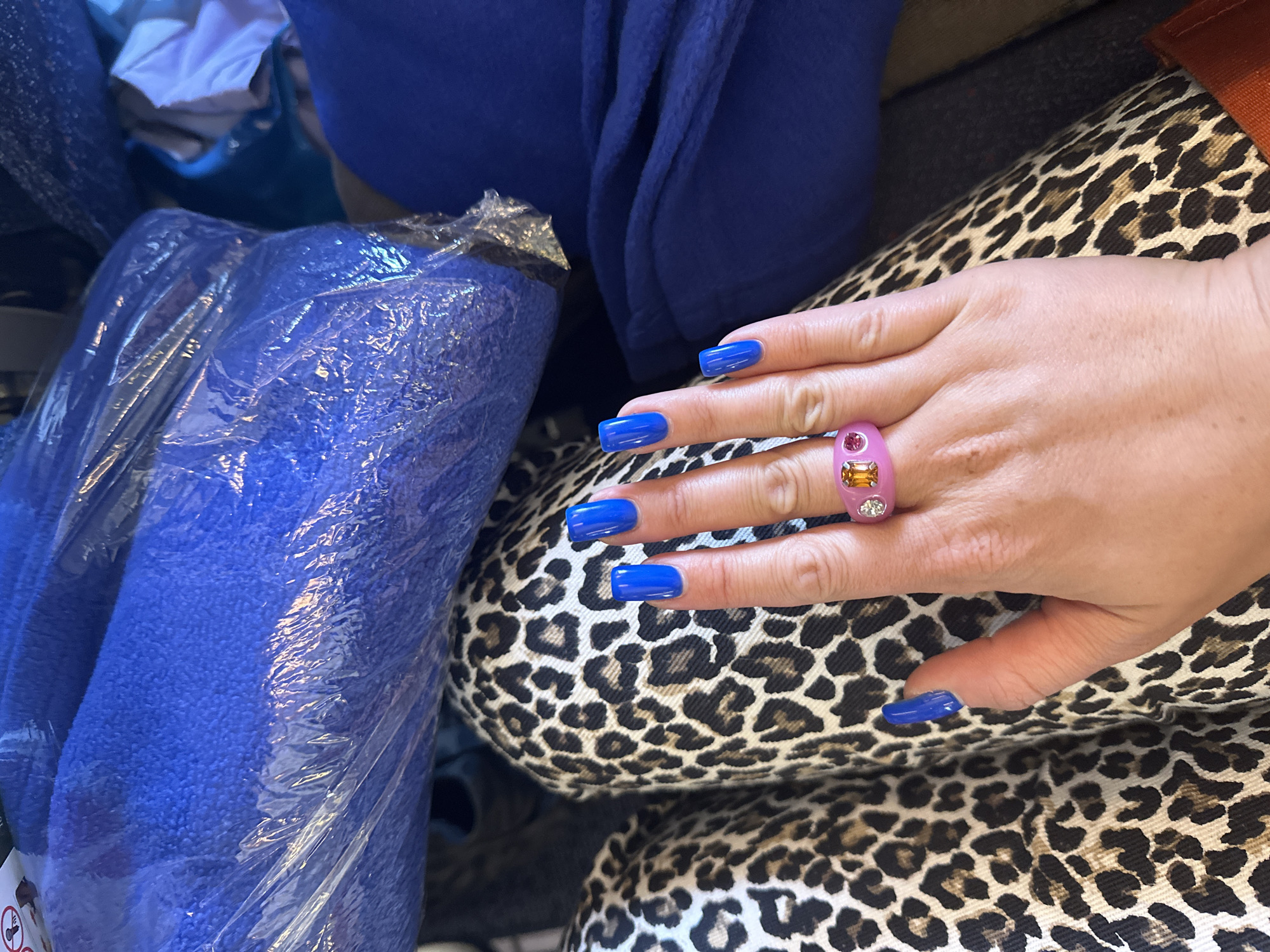 The image shows a close-up of a person's hand with bright blue nail polish and a pink ring with two stones. The hand is resting on a person's lap, which is covered in leopard-print pants. In the background, there are several folded blue blankets or fabric items, some of which are wrapped in plastic. The scene appears to be indoors, possibly in a home or a retail setting. There is no discernible information in the image that would allow for the determination of its location in Yelizovo, Russia.