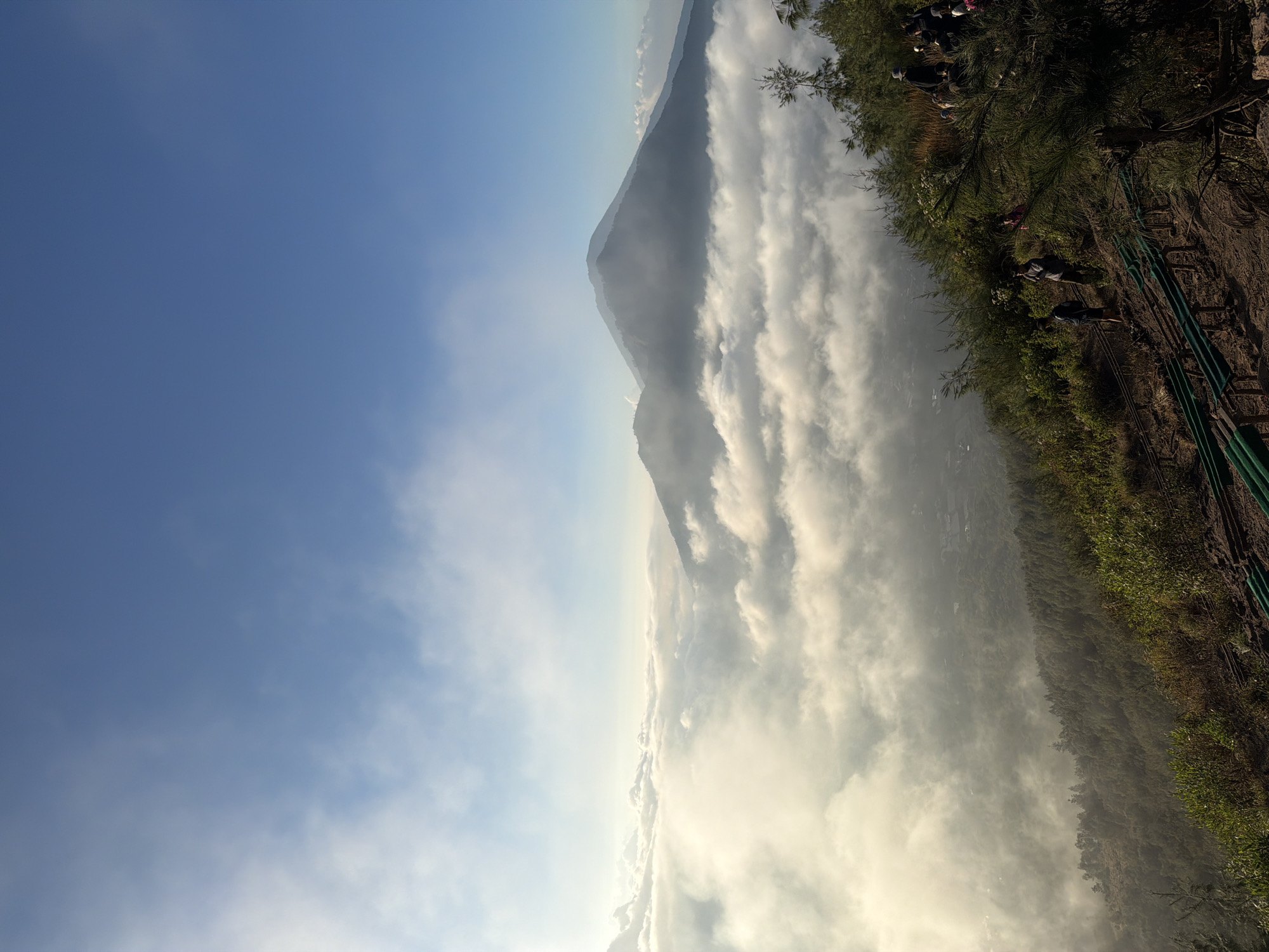The image captures a scenic mountain landscape with a bright blue sky and scattered clouds. In the foreground, a steep, vegetated slope is visible, with a dirt path winding down. Several people, dressed in casual clothing, are walking along this path, some in groups and some individually.
Further in the distance, a vast expanse of white clouds stretches across the valley, obscuring the ground below. Two prominent mountains rise above the cloud layer, with one appearing as a large, dark silhouette against the sky and the other appearing partially obscured by mist. The scene suggests a location at a high altitude, possibly Mount Batur or Mount Abang, common viewpoints in Kintamani, Indonesia, offering breathtaking views of the surrounding volcanic landscape and caldera. The lighting indicates it could be early morning or late afternoon.