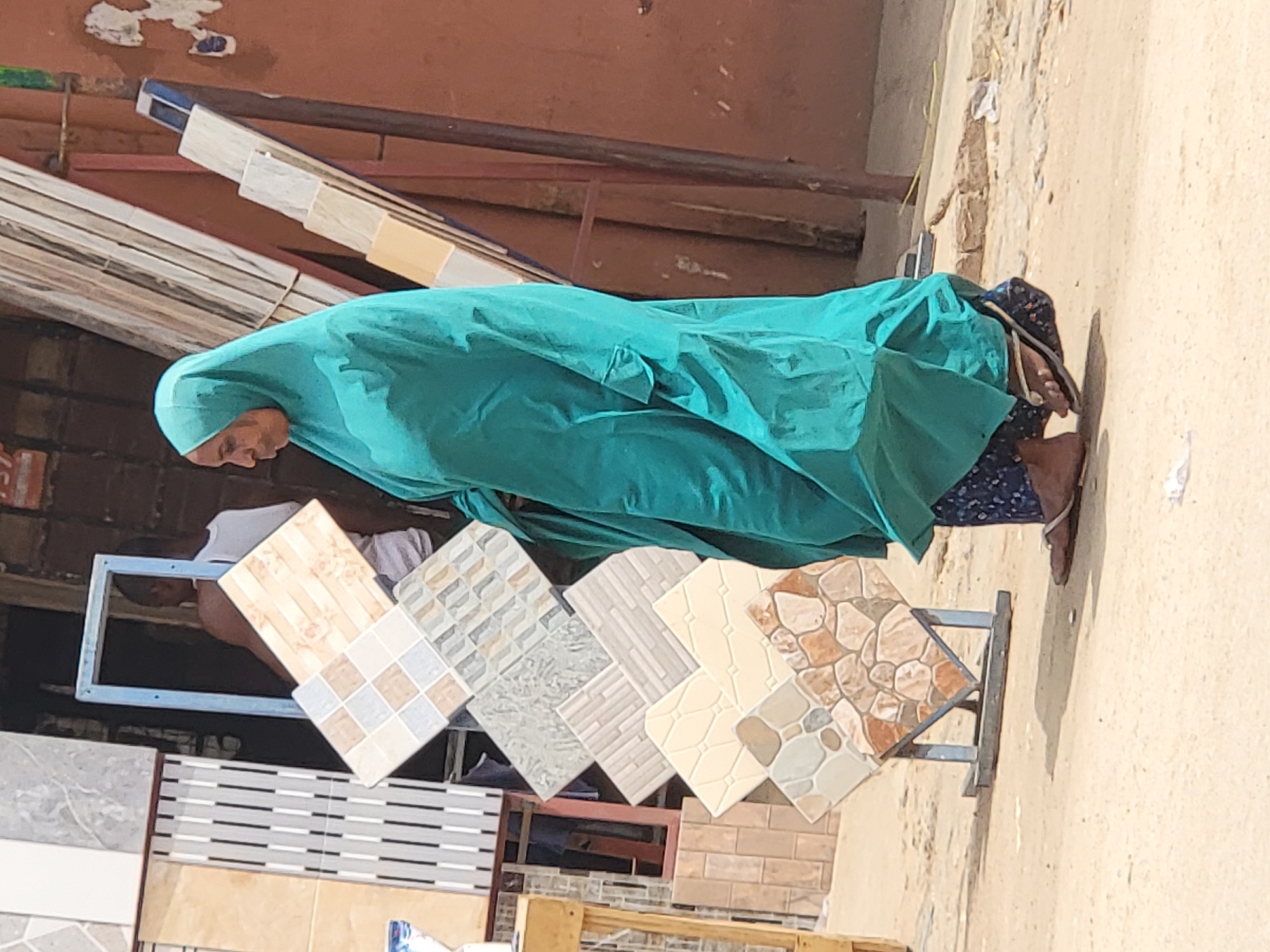 The image captures an outdoor commercial scene, likely a market stall or shop in Maiburiji, Nigeria, under bright daylight. The main subject is a person, possibly a woman, clad in a flowing teal or turquoise head covering (hijab) and a dark patterned lower garment. Their face is partially visible, with their gaze directed downwards. They are wearing sandals, and sharp shadows beneath their feet indicate strong sunlight.
In the mid-ground, a prominent display features various ceramic or floor tiles arranged on metal stands. These tiles exhibit diverse patterns and colors, including geometric squares, horizontal stripes, textured rectangles, and natural stone-like designs, in hues of beige, cream, brown, grey, and white. Another individual's arm in a white shirt is partially visible behind one of these displays.
The ground is a light, sandy, or dirt-colored surface. In the background, a reddish-brown wall or building structure is visible, showing signs of wear with peeling paint and some wooden elements. The clear, bright lighting and the outdoor setting suggest a sunny day, likely mid-day given the direct shadows. No discernible text is visible in the image.
