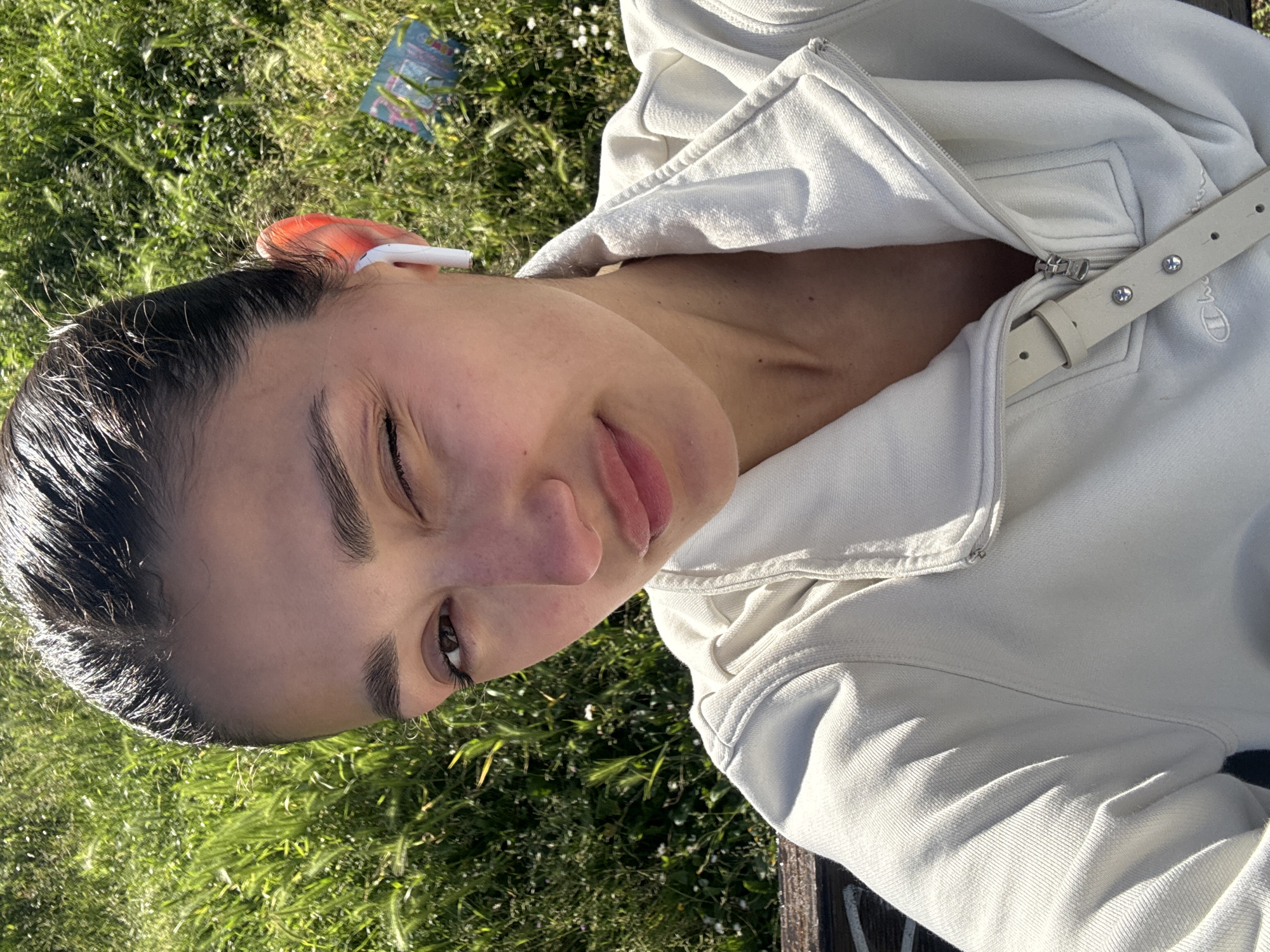 A close-up, brightly lit portrait features a young woman outdoors, her head tilted, looking towards the camera with a playful expression. Her left eye is slightly closed, and her tongue is subtly visible between her lips. Her dark hair appears slicked back or wet, framing her face.
She is wearing a light-colored (likely off-white or light grey) hooded jacket or sweatshirt, partially unzipped. A white belt with silver stud details and a visible "Champion" logo is draped over her right shoulder area. In her right ear, she wears a white wireless earbud, next to which an orange object, possibly a hair accessory or ear cuff, is visible.
The background consists of vibrant green grass, indicating a sunny outdoor setting. A small, colorful object with unreadable text is partially visible in the grass behind her head. The overall lighting suggests it is daytime, possibly with direct sunlight illuminating her face and the environment. The image conveys a relaxed and cheerful mood. The scene is set in Bar, Montenegro.