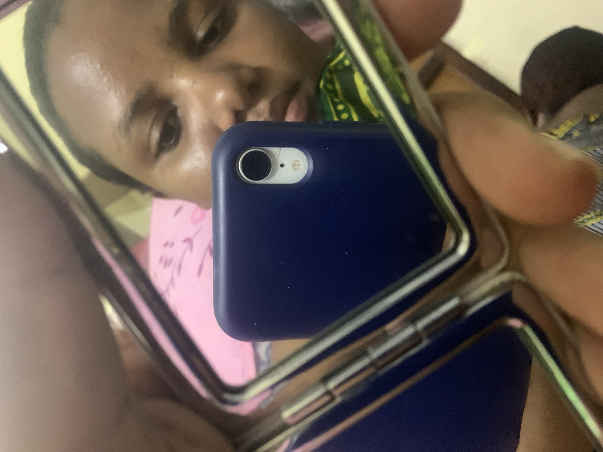A woman is taking a selfie in a mirror using her phone, the phone being blue and obscuring some of her face. The woman is wearing a green and yellow patterned top. The surroundings are out of focus, but there appears to be a pink surface behind the mirror. No information confirms the location is Dar es-Salaam, Tanzania.
