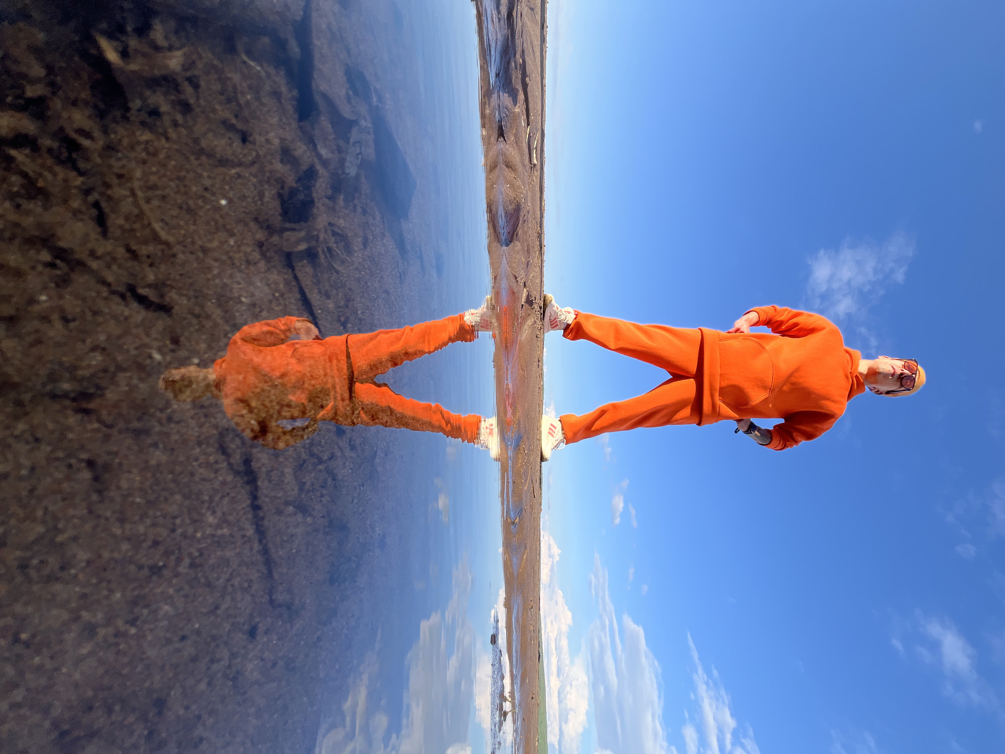 The image, taken from a low angle, presents a visually striking scene of a person and their reflection, bisected by a calm body of water. The photograph is oriented vertically, with the horizon line running through the center.
In the upper section, a person stands dressed in a bright, monochromatic orange tracksuit, comprising a hoodie and matching pants. They wear sunglasses and possibly a cap, with their head slightly tilted back and facing towards the left. White sneakers, featuring distinct stripes on the sides, are visible on their feet. This half of the image is dominated by a clear, vibrant blue sky dotted with sparse white clouds, suggesting a bright and sunny day.
The lower section reveals a near-perfect, mirror-like reflection of the person and the sky in the still water. The reflection of the person in the orange tracksuit, with their distinctive white sneakers, is precise and sharp, appearing as if another individual is standing inverted below them. The water also clearly mirrors the blue sky and clouds. The narrow strip of land or shore on which the person stands, along with the visible ground beneath the shallow water, is dark and textured.
The activity depicts the person posing for a photograph, strategically using the highly reflective water surface to create an artistic and symmetrical composition. The time of day is clearly daytime due to the bright light. Notable details include the vivid orange of the attire, the pristine clarity of the reflection, and the serene quality of the water. No specific legible text is visible, apart from the prominent stripes on the sneakers. The scene captures a tranquil moment outdoors, likely on the shore of a lake or large puddle in Chaikovsky, Russia.