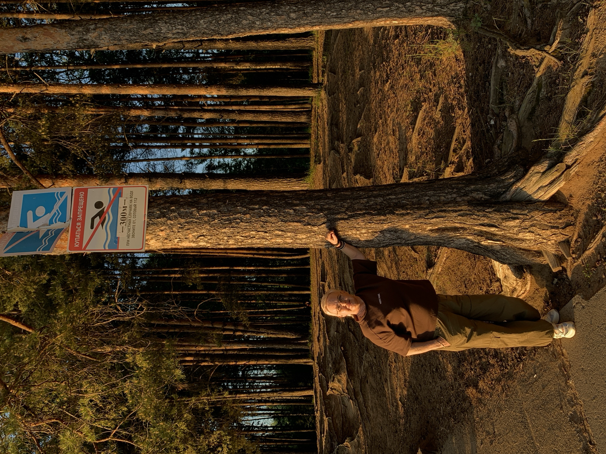 The image captures a man standing by a tree in a forest in Chaikovsky, Russia. He is wearing a brown t-shirt, olive-green pants, and white sneakers. His left arm is resting on the tree trunk. Next to the tree is a sign reading, "КУПАТЬСЯ ЗАПРЕЩЕНО" which translates to "Swimming is Prohibited". The sign also states, "-300м- При несчастных случаях на воде звоните 01, сотовый 112" which translates to "-300m- In case of accidents on the water, call 01, mobile 112". The background reveals a forest with tall, slender trees, and the ground near the man appears to be covered in dirt and tree roots. The light suggests it is either early morning or late afternoon.