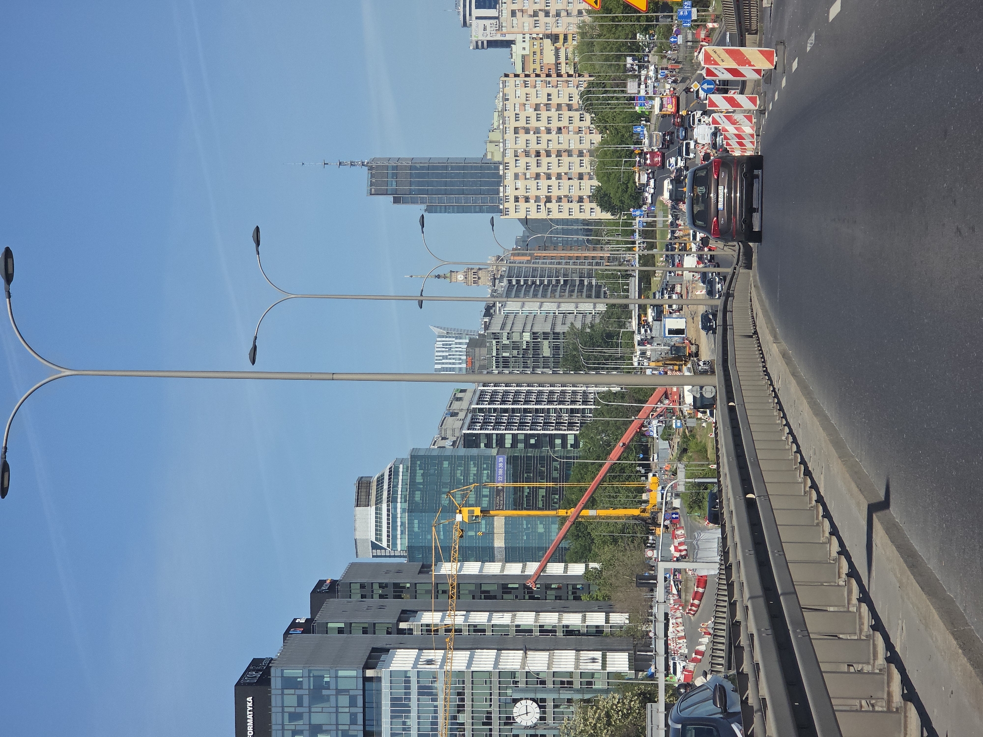 The image depicts a street scene in Warsaw, Poland, likely taken from a bridge or elevated roadway. The focus is on the urban landscape, including buildings and traffic. The foreground features the side of a road with a grey metal barrier. Beyond the barrier, there is construction work and various traffic signs. A line of traffic is visible, including several cars, a bus, and trucks. The background showcases the Warsaw skyline, consisting of a mix of modern high-rise buildings and older architectural styles. A clear blue sky fills the upper portion of the image. Several tall street lights are prominent on the left side of the image.