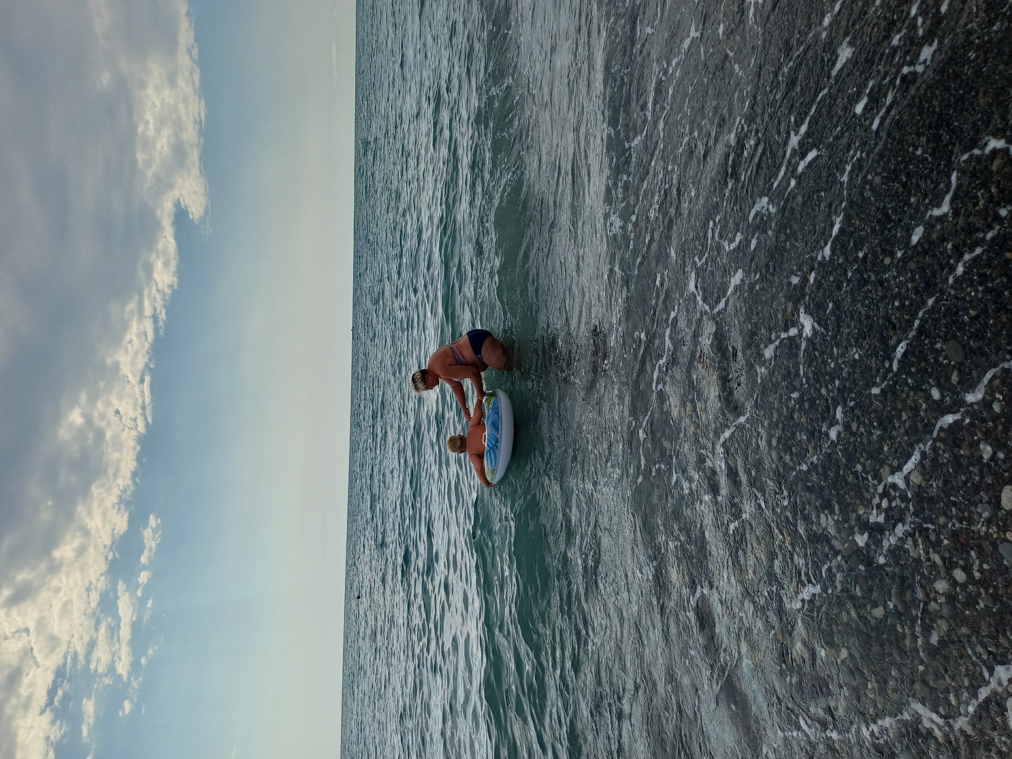 This is a vertical, eye-level, full shot of two people, a woman and a child, in the water at a pebble beach. The woman, wearing a dark bikini, is bent over in the water, holding onto a child who is sitting on an inflatable ring. The child is facing away from the camera and is wearing light-colored clothing. The water is a dark, wavy, and translucent blue. The seabed is visible through the water and is covered in dark pebbles. The sky is a light blue with fluffy white clouds and is visible in the upper left quadrant of the image. The sun appears to be high in the sky, suggesting it is daytime. The scene appears to be a leisurely day at the beach in Sochi, Russia.