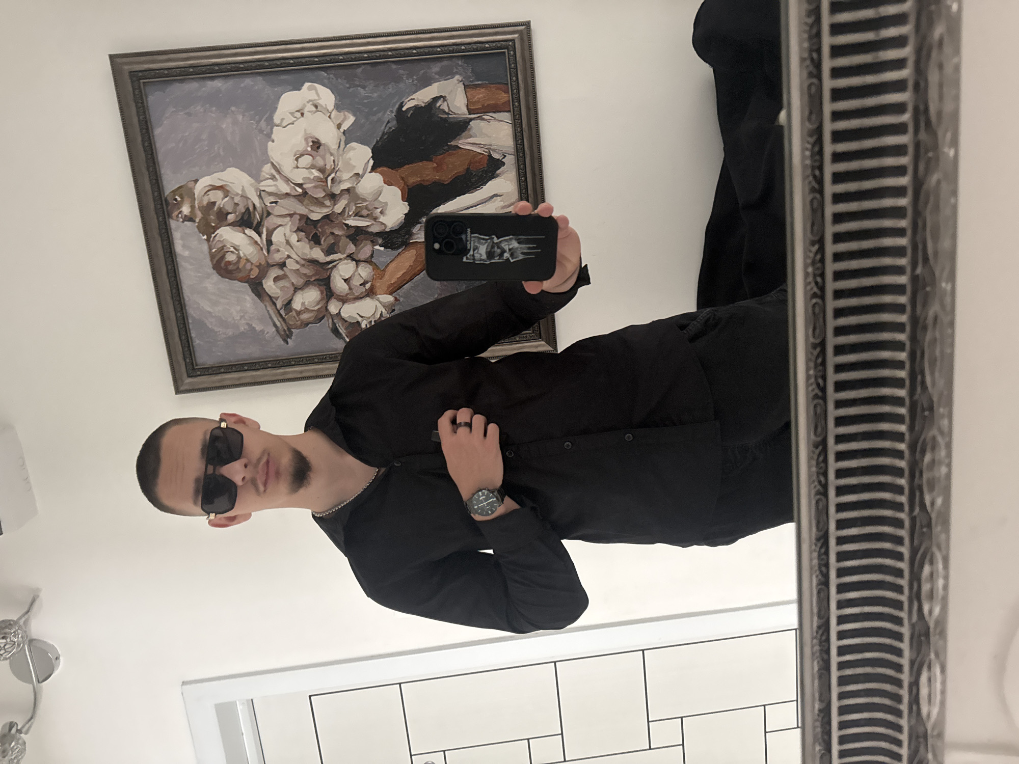 A man is taking a selfie in a mirror. He is wearing a black shirt, black pants, and sunglasses. He has a watch on his left wrist. The man is holding a black smartphone with a picture on the case. Behind the man, there is a framed painting with flowers. Below him, there is a white tiled wall. The location is not discernible from the image. No information suggests this location is in the city of Pazardzhik, Bulgaria.