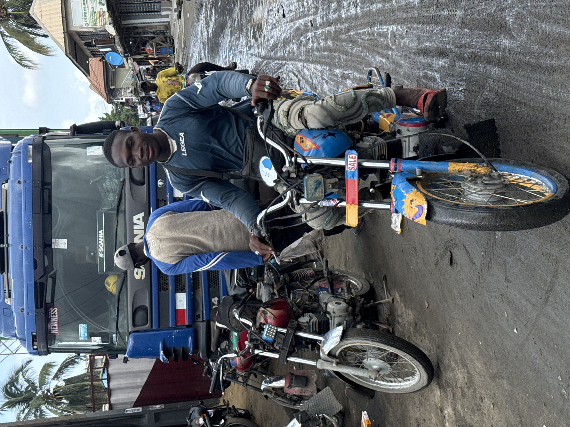 This portrait-oriented image captures a bustling street scene in Lekki, Nigeria, centered around a young man on a customized motorcycle, looking directly at the viewer.
The primary subject, a man with dark skin and short dark hair, wears a dark blue long-sleeved shirt with the "LEGEA" logo visible on the right chest. He has rings on his fingers and is sporting light-colored, possibly homemade, knee pads. He sits astride a distinctive motorcycle with a blue frame and yellow painted accents on the tank and fenders. A small, colorful sign reading "SALE" is attached near the handlebars.
Behind the main subject, another person is visible on a different motorcycle, wearing a grey and blue top. To the left, a large blue Scania truck is prominent, with "SCANIA" clearly visible on its grille and "NEWNESS" written on its bumper. Other people are sparsely scattered in the background, including one person in a bright yellow shirt further down the street. The ground appears to be a mix of unpaved earth and possibly wet, grey asphalt, suggesting recent rain or damp conditions.
In the background, there are commercial buildings with various signs, some illegible, and a palm tree is visible on the left, indicative of a tropical setting. The sky is overcast, providing soft, diffused daylight. The scene portrays a lively urban or semi-urban environment, characteristic of a marketplace or busy road where motorcycles are a common mode of transport.
