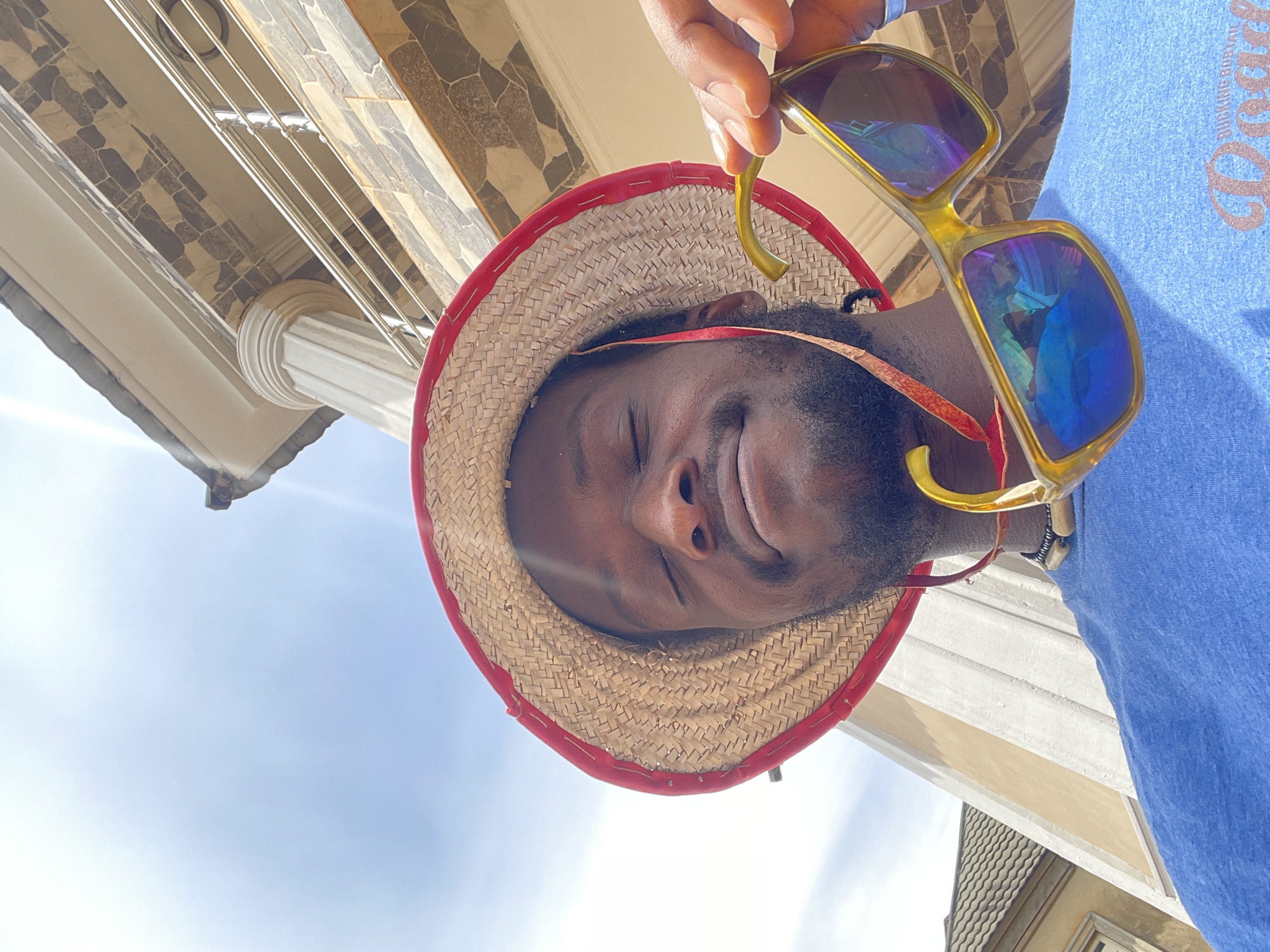 A man with a straw hat and sunglasses is photographed from a low angle on a sunny day, likely in a residential area of Abuja, Nigeria. The man is wearing a straw hat with a red trim, and he is holding a pair of sunglasses with yellow frames. The sunglasses have blue-tinted lenses that reflect the sky and surroundings. He is wearing a blue t-shirt. The backdrop includes a building with stone accents, a white pillar, and a glimpse of a tiled roof. The sky is visible in the background.