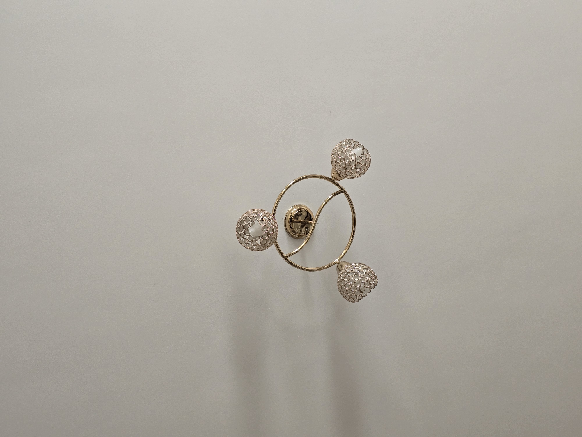 This media file shows a close-up, top-down view of a modern ceiling light fixture. The fixture is made of gold-toned metal and has a circular base from which three arms extend. Each arm terminates in a spherical lampshade made of a delicate, textured material that appears to be woven or composed of small, reflective beads, creating a glittering effect. Inside each lampshade, a light bulb is visible, appearing to be off. The overall design is contemporary and decorative, suggesting it is intended to be a stylish accent piece in a room. The background is a plain, off-white ceiling, providing no additional environmental context. There is no visible text in the image. The lighting suggests it is indoors, likely during daytime, as there are soft shadows cast on the ceiling from the fixture, indicating ambient light.