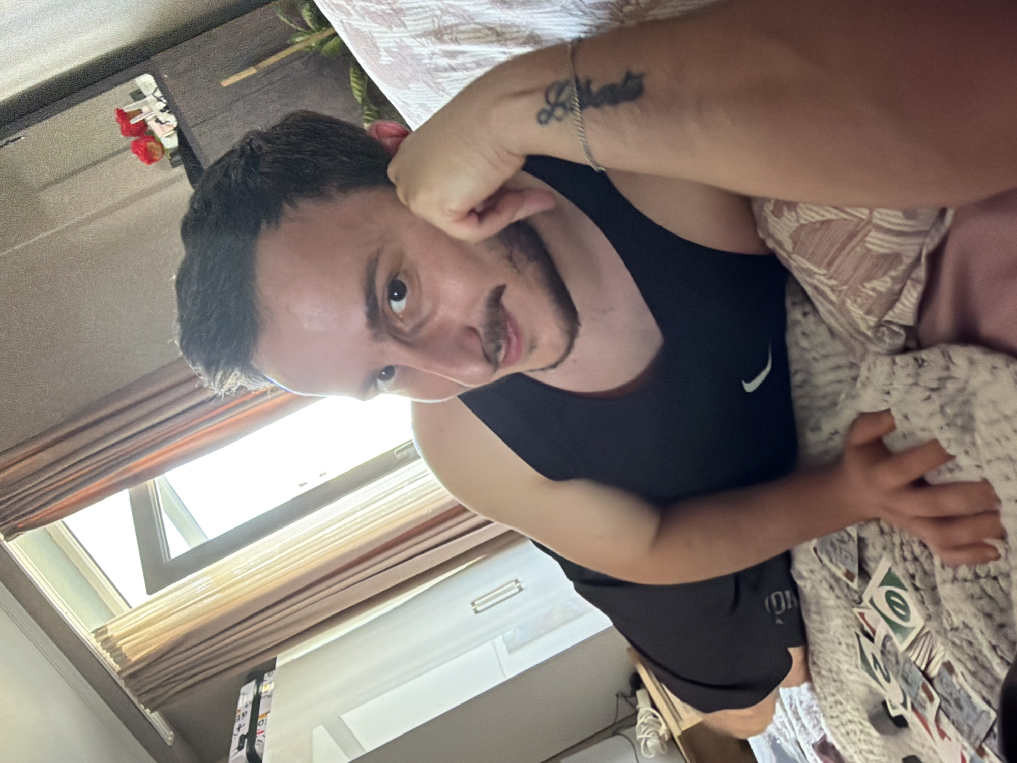 A young man with short, dark hair and a mustache lies on a bed, propping his head on his hand. He is wearing a black tank top and black shorts. The bed is covered with a textured blanket and some UNO cards are scattered on it. Behind him, a window with light brown curtains is visible, and to the left, a white wardrobe with handles can be seen. A shelf with a small decorative item, possibly flowers, is mounted on the wall above the wardrobe. The overall environment suggests an indoor setting, likely a bedroom. There is no information to suggest the location is Tbilisi, Georgia.