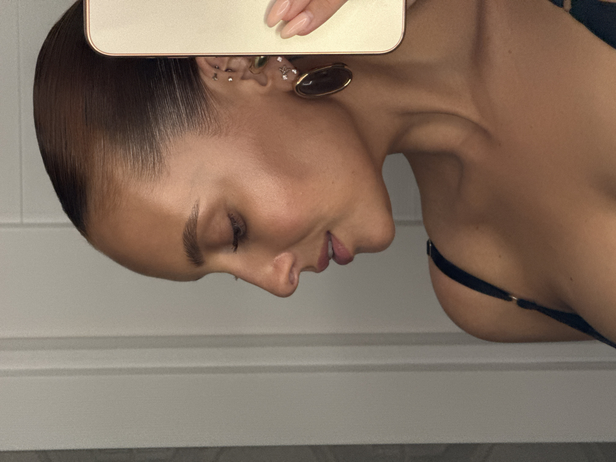 In a close-up, profile view, a woman with her hair slicked back is holding a gold-colored smartphone to take a selfie. She is wearing large, dark oval earrings and multiple smaller piercings in her ear. Her right hand, with manicured nails, is visible holding the phone. The background appears to be an interior wall with vertical paneling. The image does not provide any information about the location in the city of Chaikovsky, Russia.