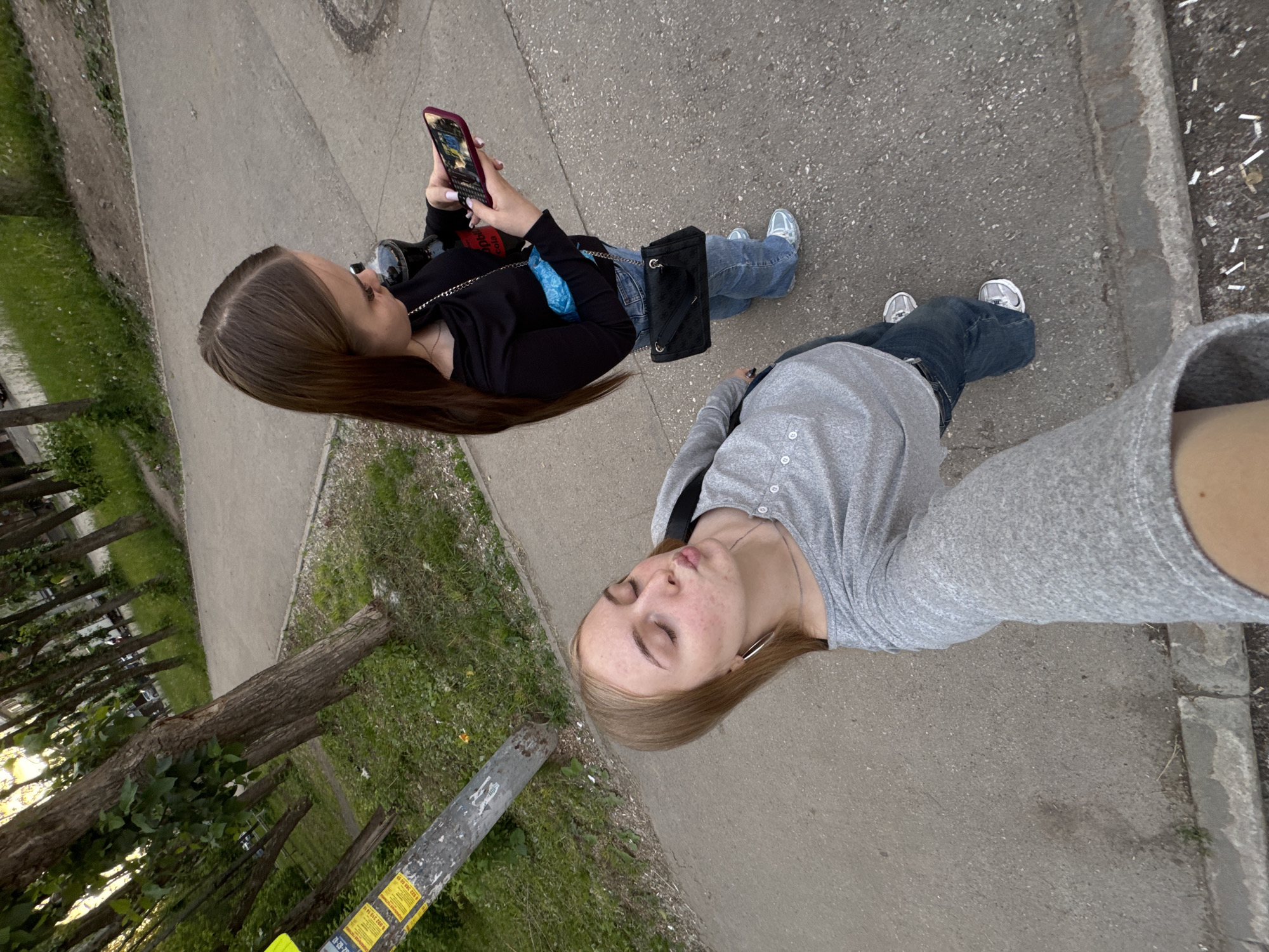Two young women are depicted outdoors on a paved asphalt path in Perm, Russia, captured in a high-angle selfie. The woman in the foreground, with light brown hair parted on the side, has her eyes closed and lips pursed. She wears a grey long-sleeved Henley-style shirt, blue jeans, silver hoop earrings, and a thin silver chain necklace. Her right forearm, holding the camera, is visible at the bottom. Mild facial blemishes are present.
The second woman, standing behind and to the right, has long dark brown hair parted in the middle. She is observed looking down at a red smartphone, which displays a screen with what appears to be a keypad. She wears a black long-sleeved top with a draped neckline, light blue wide-leg jeans, and white sneakers with light blue accents. A black shoulder bag with a silver chain strap hangs from her left shoulder, and a light blue pouch is tucked into her waistband. A dark bottle, featuring a red label and possibly containing Coca-Cola, is held near her hip.
The setting is an outdoor area featuring a grey asphalt path. To the left, a grey utility pole displays multiple yellow rectangular advertisements with Russian text, including phone numbers (e.g., "8 922 388 50 95", "8 982 715 28 64"). The path is bordered by a concrete curb, adjacent to grassy patches with scattered trees featuring green foliage. Small white paper fragments are visible on the ground near the curb in the bottom right. The lighting is consistent with an overcast day or late afternoon.