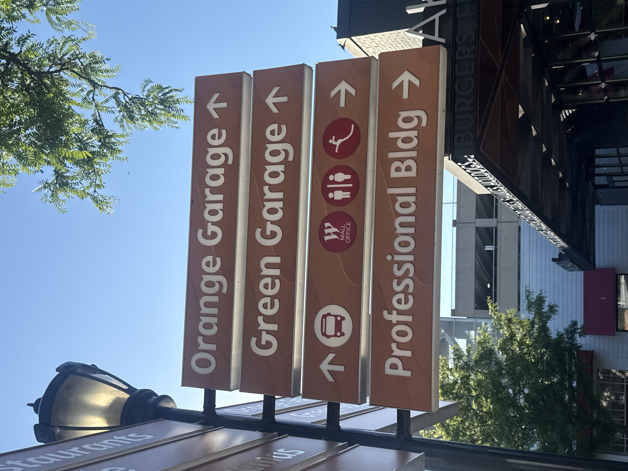 The image shows a direction sign in Skokie, Illinois. The sign has four vertical panels pointing to the right. The first panel says "Orange Garage" with an arrow pointing upward. The second panel says "Green Garage" with an arrow pointing upward. The third panel has three symbols: a person skiing, a man and woman symbol, and the Westfield Old Orchard Mall Office logo. Under the logo is a bus symbol and an arrow pointing up and left. The fourth panel says "Professional Bldg" with an arrow pointing right. Above the sign, a building has a sign that reads "BURGERS F". A tree and a light post are partially visible in the shot.