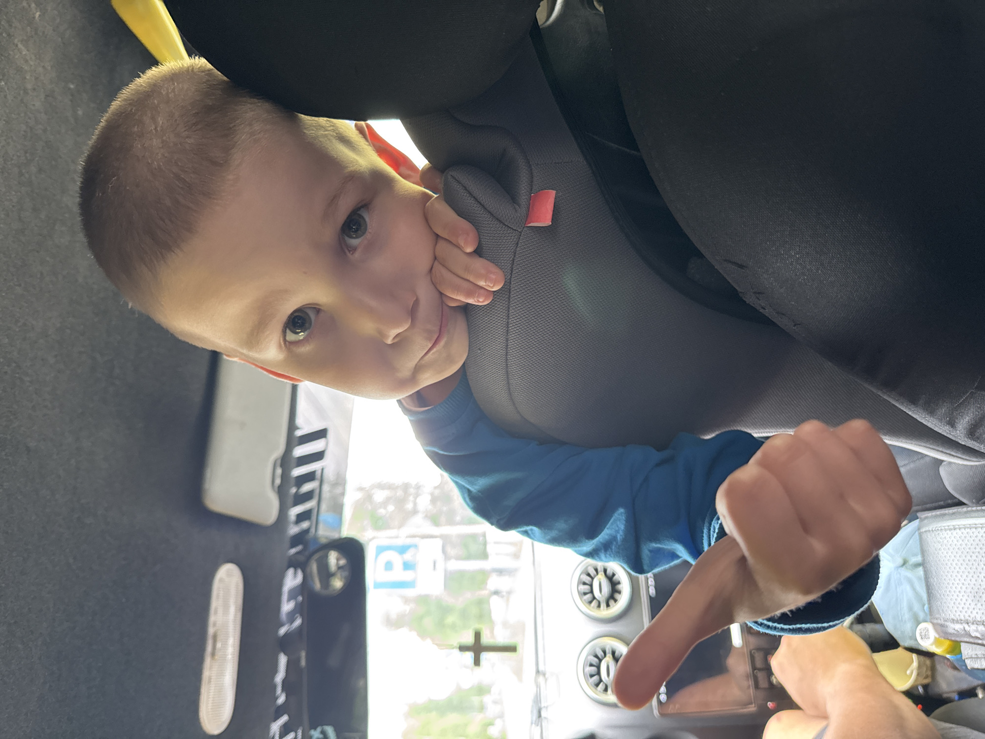 A young boy with short brown hair, looking towards the camera, rests his head on a car seat. He is wearing a blue long-sleeved shirt and gives a thumbs-up gesture with his left hand. The background shows the interior of a car, including a dashboard with circular vents, a rearview mirror, and a cross hanging from the windshield. Outside the car, a blue sign with the letter "P" is visible, along with trees and buildings, suggesting an urban or suburban environment, possibly in Chaikovsky, Russia.
