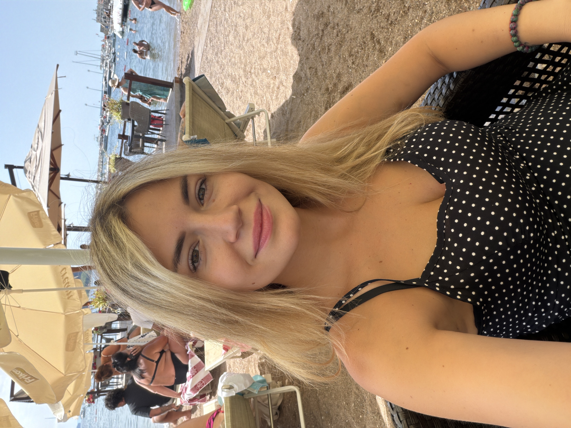 The image captures a woman with blonde hair, wearing a black and white polka dot dress, sitting at an outdoor cafe on a beach. The cafe appears to be located on the sand, with beach umbrellas providing shade. In the background, the sea is visible with several boats docked, and other people are enjoying themselves on the beach and in the water. The scene suggests a relaxed atmosphere typical of a summer day at a coastal resort. The location is the city of Budva, Montenegro, a popular tourist destination known for its beaches and historical old town.