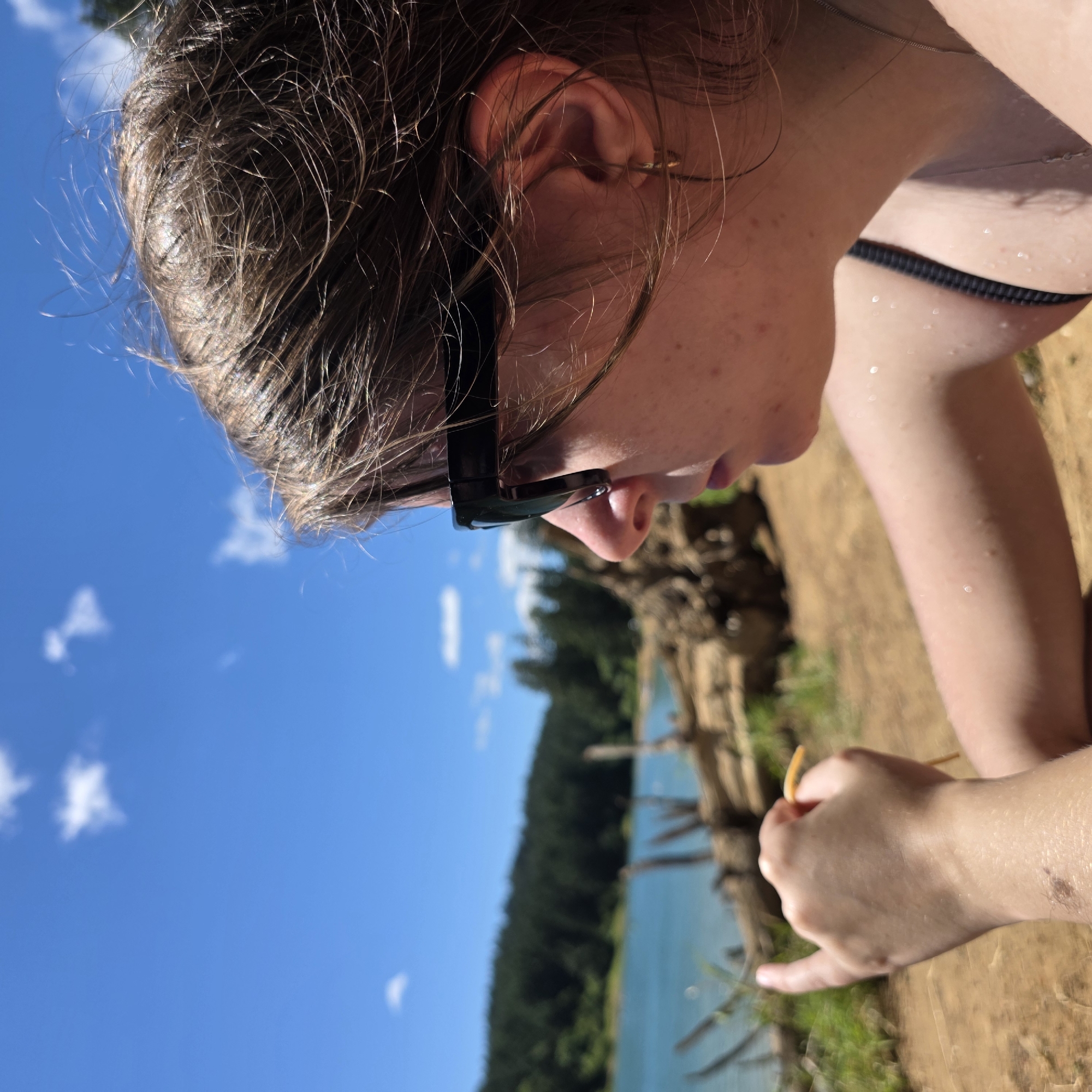 A medium shot shows a person wearing black sunglasses and a black bikini top, looking out at a blue lake bordered by a dark green forest under a bright blue sky with scattered white clouds. The person's right hand is in the foreground, holding a yellow object. The person's skin is wet, with small water droplets visible on their arm and neck. The location is Cherry Grove, United States.