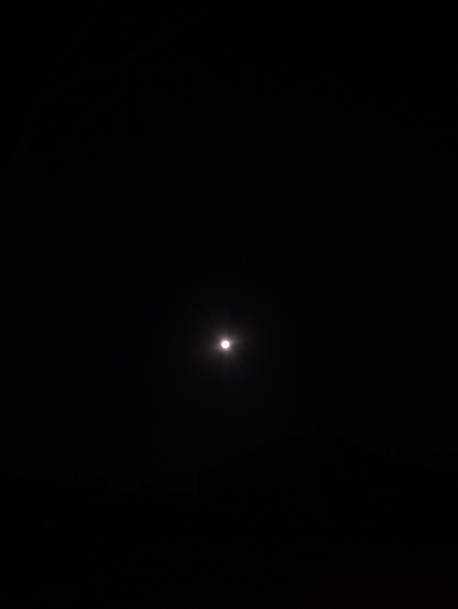 A single, bright, circular light source, likely the moon, is visible in a completely dark sky. Below the celestial body, a faint, dark silhouette of a landform, possibly hills or mountains, is discernable against the slightly lighter horizon. The overall image conveys a scene of a clear night with minimal light pollution, allowing the moon to be a dominant feature. The location in Kalisongo, Indonesia, suggests a rural or semi-rural setting where such unobstructed night skies are more common. There are no people or specific objects identifiable in the scene, only the natural elements of sky and land under the illumination of the moon.