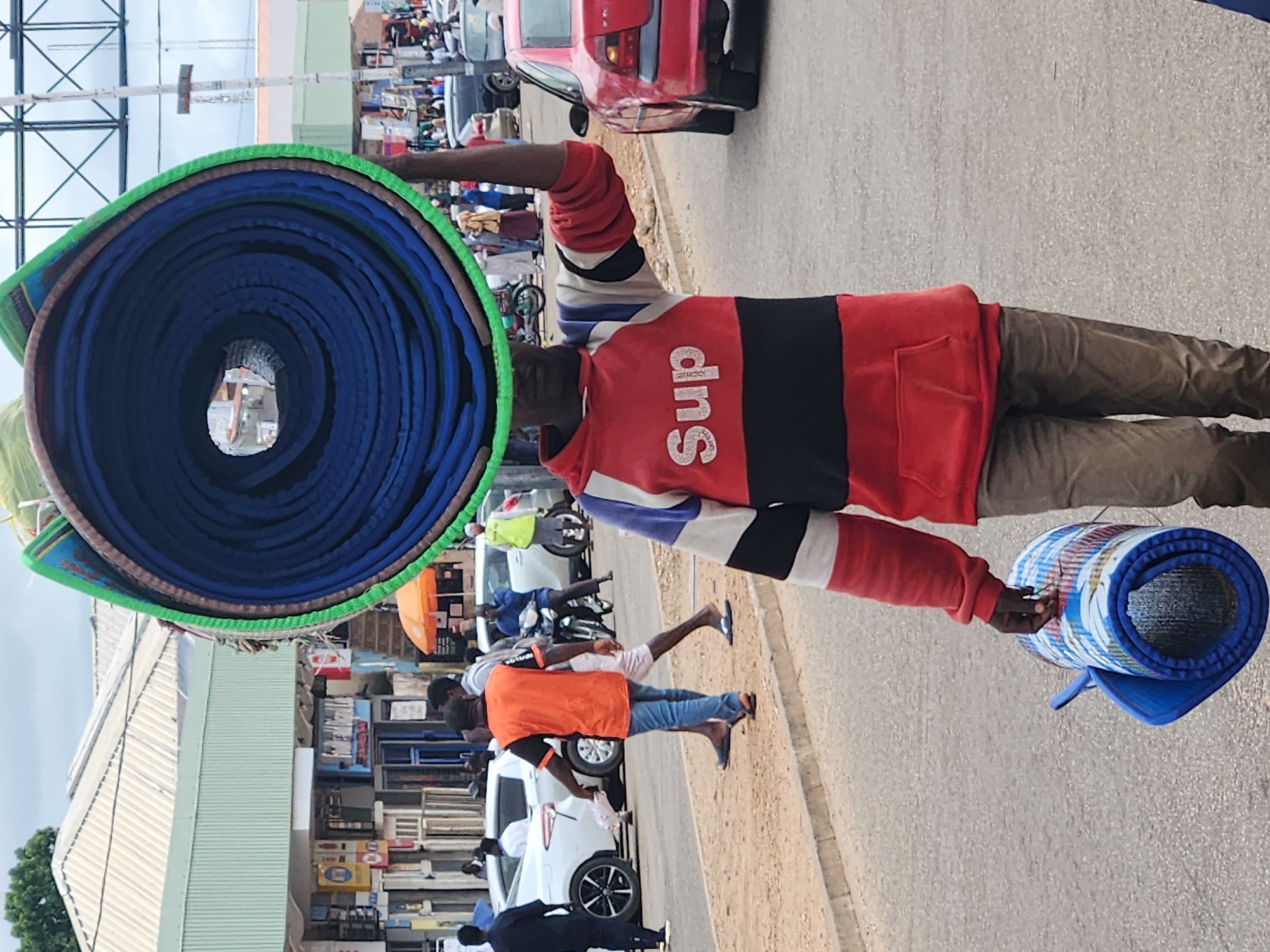 A man is walking down a street carrying a large rolled-up mat on his head and another rolled-up mat in his hand. He is wearing a red hooded sweatshirt with the word "Sup" printed on it in white letters. He is also wearing brown pants. The street is paved and there are buildings on either side. There are other people and vehicles in the background. The sky is cloudy.