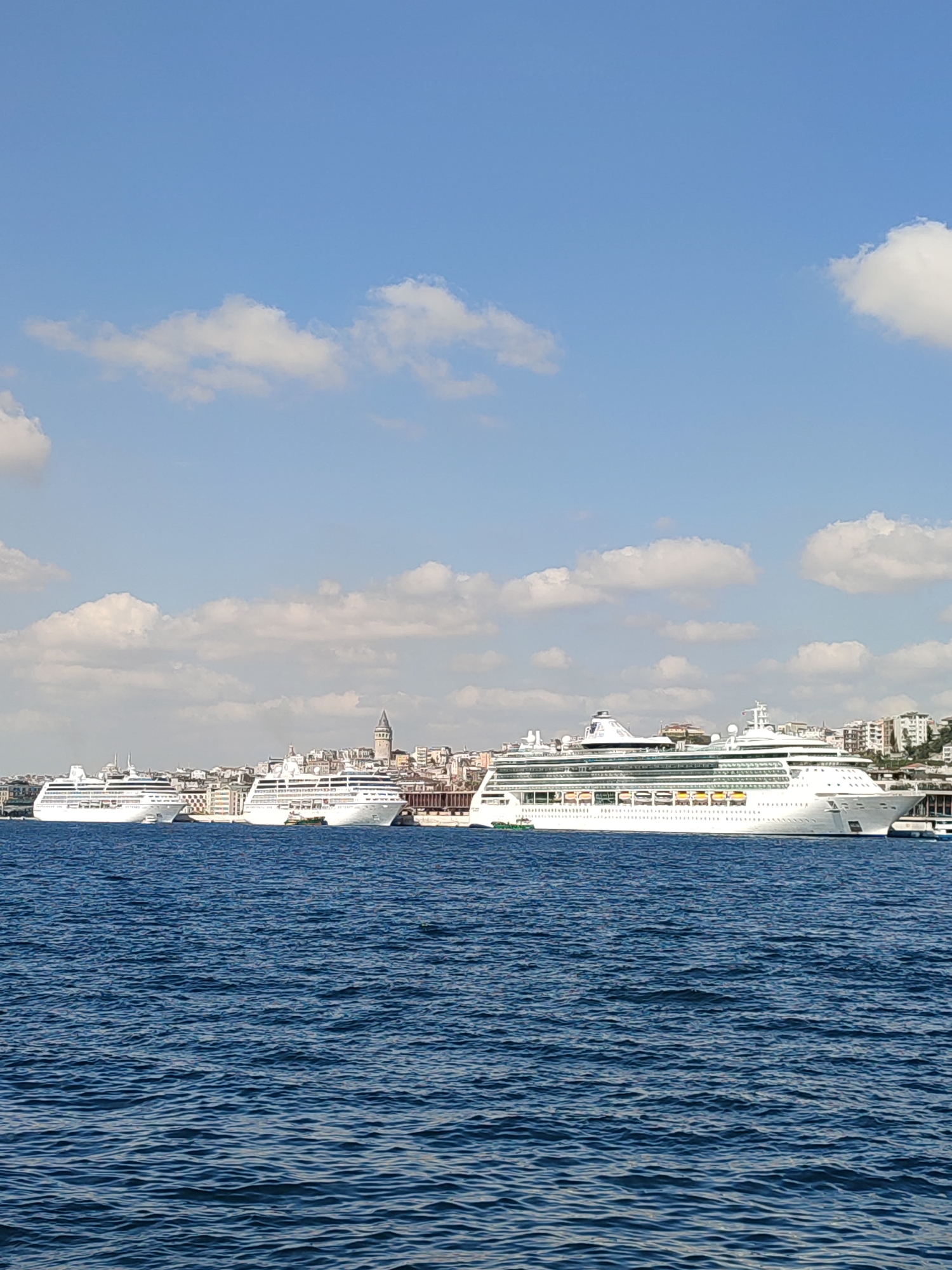 The image shows a panoramic view of cruise ships docked along the waterfront of Beyoğlu, Turkey. In the foreground, the deep blue water of the Bosphorus is visible, with small ripples on its surface. Three large, white cruise ships are positioned parallel to the shore. The largest ship, in the middle right of the frame, has multiple decks and rows of windows. The other two ships are located to its left, with the furthest one partially obscured by the middle ship.
Behind the cruise ships, the skyline of Beyoğlu features a dense collection of buildings, ranging in height and architectural style. A prominent historical structure, the Galata Tower, stands tall amidst the urban landscape. The buildings appear to be a mix of residential, commercial, and possibly administrative structures. The shoreline is lined with piers and what appears to be a port or harbor area. A bridge structure is visible on the right side of the image.
The sky above is a clear, bright blue, dotted with scattered white, fluffy clouds. The lighting suggests it is daytime, likely late morning or early afternoon, with the sun illuminating the scene and casting light on the water and the ships. No people are discernible in the image, and the scene depicts a static, peaceful maritime environment. The overall impression is one of a busy port city with significant maritime traffic.