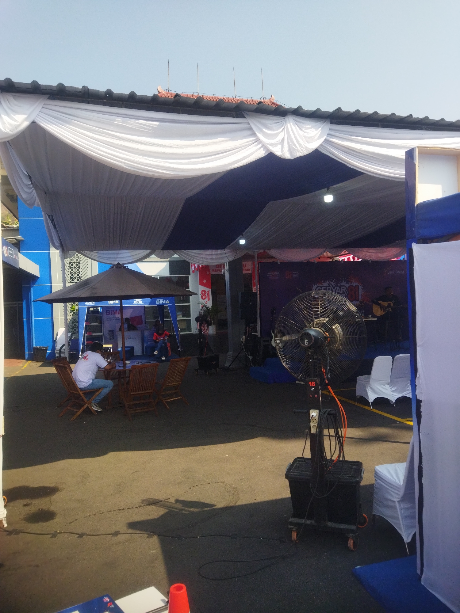 The image depicts an outdoor event setup under a large tent. In the foreground, a person wearing a white shirt and blue jeans sits at a wooden table with four chairs, under a dark umbrella. The table and chairs are arranged on an asphalt surface. In the midground, a large industrial fan with an orange cord is prominently placed on a wheeled black cart. Behind the fan, a stage is set up with a musician playing an acoustic guitar and another person standing nearby. There are two white chairs covered with white fabric placed in front of the stage. The background features a blue building with windows and signage. The overall environment appears to be a public space, likely in the city of Giripurwo, Indonesia, given the context provided. The lighting suggests it is daytime, with clear skies.