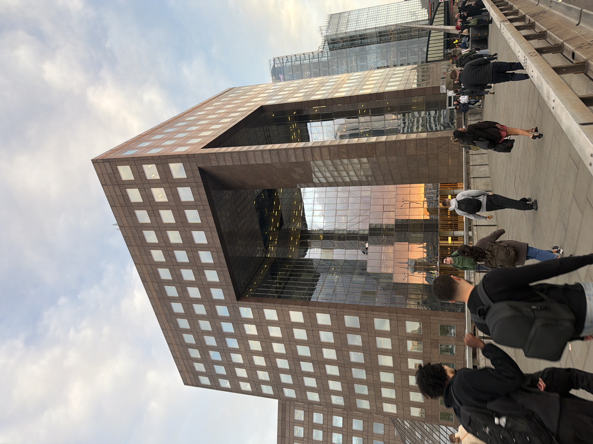 The image captures a view of the city of London, United Kingdom. In the foreground, there is a unique building with a large opening or archway in the middle section. The building has a grid-like pattern of windows on its exterior. A crowd of people is walking along a pedestrian walkway, possibly a bridge, in front of the building. The people are dressed in casual clothing and appear to be engaged in pedestrian traffic. In the background, there are modern glass-and-steel skyscrapers that are reflective, showing a cityscape. The sky is overcast with muted light, and the overall scene has an urban, contemporary feel.