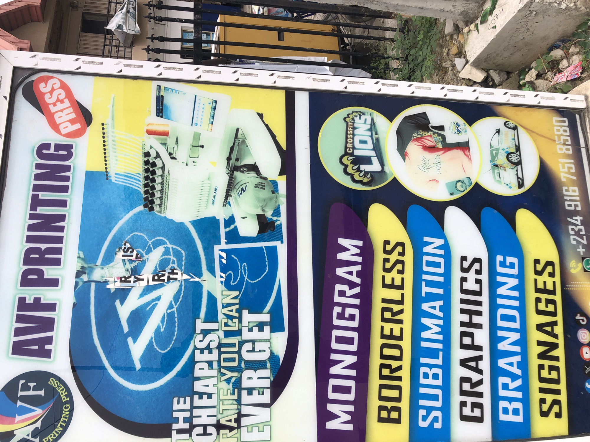 The image shows a sign for AVF Printing, advertising "the cheapest rate you can ever get," and promoting services like monogramming, borderless printing, sublimation, graphics, branding, and signage. It features images of printing equipment, logos like "Crossfit Lions," and social media icons. The phone number +234 916 751 8580 is also displayed. The sign appears to be located outdoors, possibly in the city of Ado, Nigeria, based on the phone number's country code.