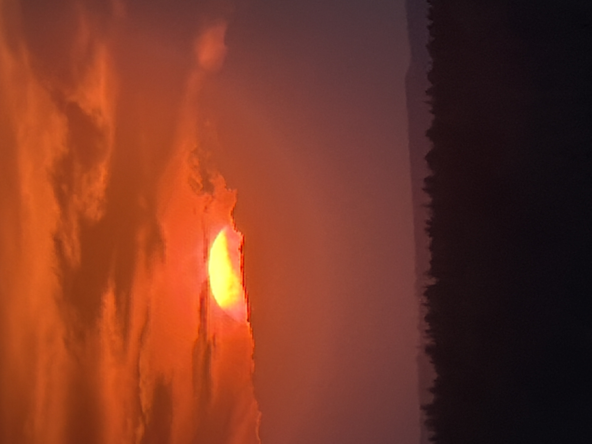The image depicts a sunset in a smoky environment, possibly due to wildfire smoke. The sun is obscured by orange-tinted clouds and haze. To the right of the frame, a dark silhouette of what appears to be a treeline is visible. The overall scene creates an impression of a hazy, potentially unhealthy atmospheric condition. No people are visible. The location is unknown.
