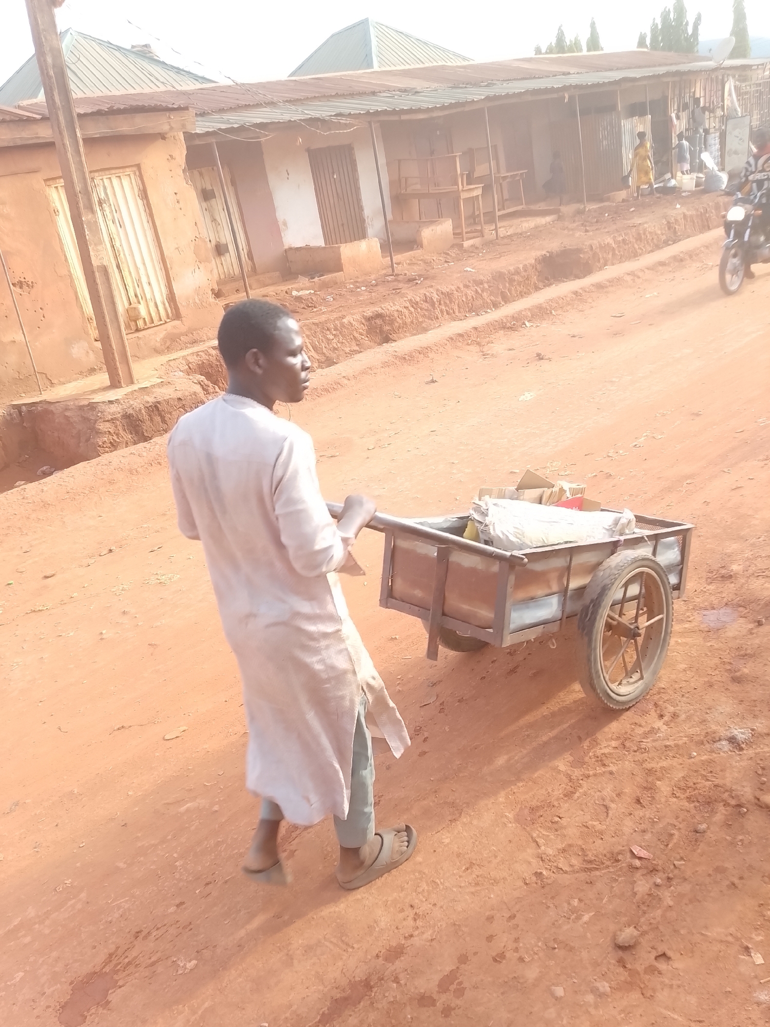 The photograph captures a street scene in what appears to be a semi-urban or rural area of Yelwa, Nigeria. The central subject is a man, seen from behind and slightly to his right, actively pushing a two-wheeled metal handcart along an unpaved, reddish-brown dirt road. The cart is loaded with various items, including a large white sack and cardboard boxes. The man is dressed in a light-colored, long-sleeved tunic over light blue trousers and wears sandals, his head turned to the right as he concentrates on his task.
The setting consists of a dusty, uneven dirt road, flanked by raised sections of earth or ditches. Along the roadside, a series of single-story buildings with corrugated metal roofs are visible. These structures have walls of varying finishes, some appearing to be mud or concrete, with visible metal doors or gates. Outside one of the buildings in the background, several pieces of wooden furniture, possibly tables or shelves, are displayed, suggesting a local shop or stall.
In the background, other people are visible, contributing to the sense of daily life. A person in a yellow dress stands near one of the buildings, and further down the road, a motorcycle with a rider is partially captured on the far right. The time of day appears to be late morning or early afternoon, indicated by the bright sunlight and visible shadows, suggesting clear and likely warm weather. There is no discernible text visible in the image. The scene portrays a common mode of transport and commerce in a local community.
