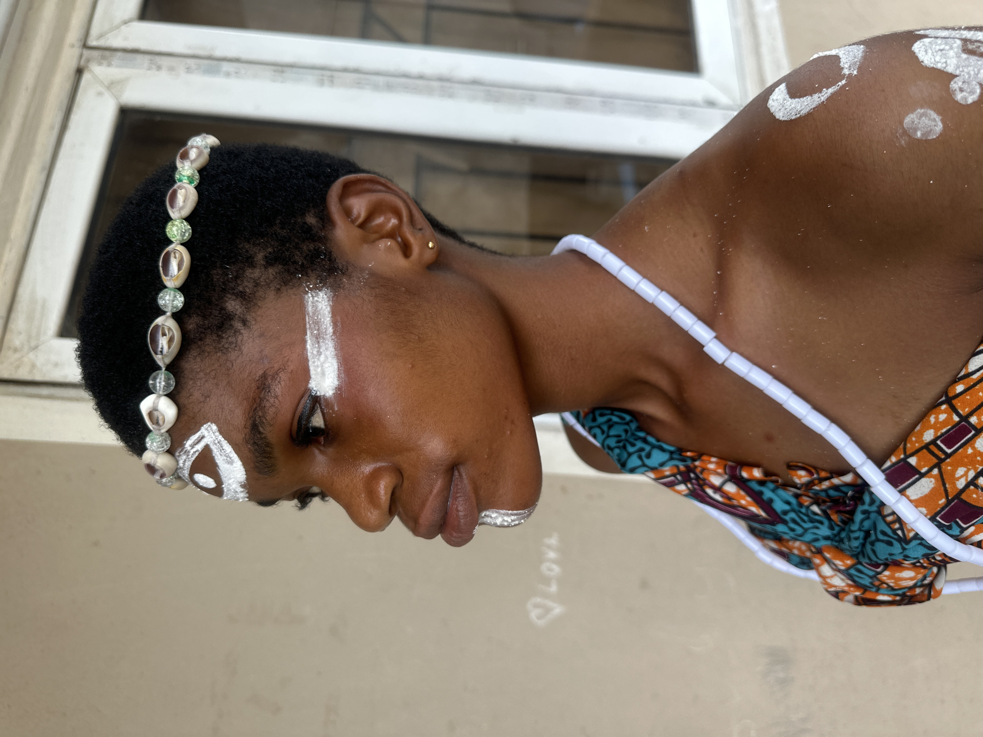 The image shows a young Black woman adorned with traditional face and body paint, and shell and bead accessories. She wears a headband made of shells and beads, a beaded necklace, and an outfit with vibrant, geometric patterns. White paint is applied in decorative patterns on her forehead, cheeks, chin, and shoulder. She is positioned in a profile view, looking to the left. In the background, a beige wall with a chalk drawing of a heart and the word "LOVE" is visible, along with a white-framed window. The context places this scene in the city of Badagry, Nigeria, a historic coastal town known for its cultural heritage.