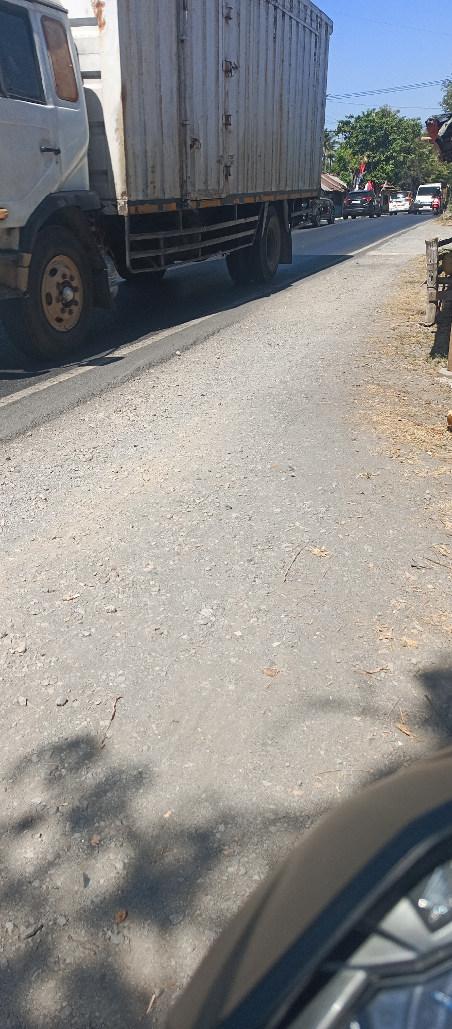 The image depicts a road in Arjasa, Indonesia, on a sunny day. A large white cargo truck is prominently featured on the left side of the frame, positioned on a paved road with visible gravel. The truck is stationary or moving slowly. In the background, other vehicles, including a black SUV, a white van, and a red scooter, are visible on the road, suggesting traffic. Trees and some structures with corrugated roofs line the side of the road. Shadows of foliage are cast on the road surface, indicating the presence of trees. The foreground shows the edge of a vehicle, possibly a motorcycle, with its headlight reflecting light. The overall scene appears to be a typical street environment in a town or rural area in Indonesia.