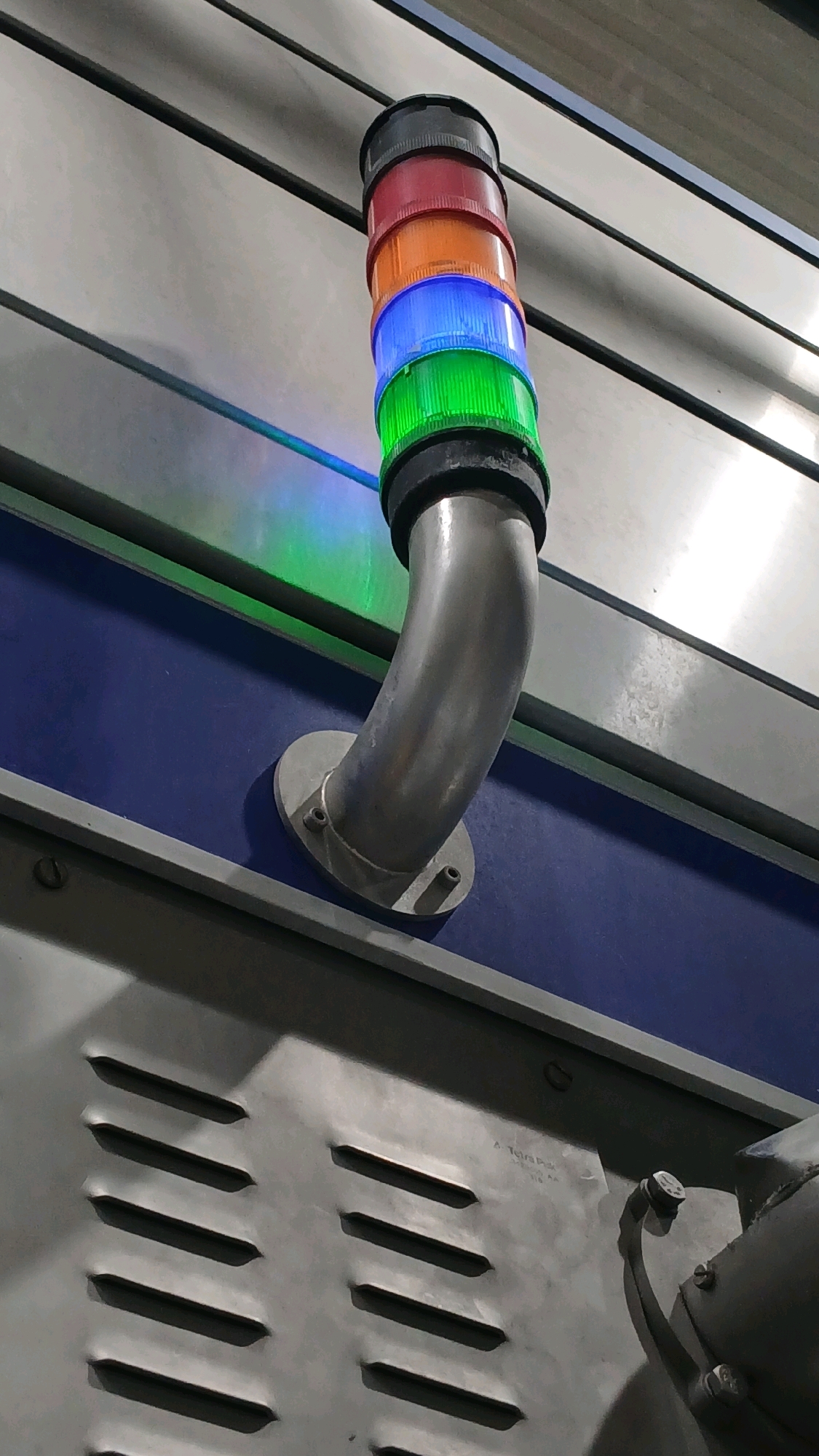 A stack light with red, orange, blue, and green lights is attached to a metallic pipe that curves out from a blue panel. The stack light is illuminated, with the green light casting a reflection on a metallic surface. The background consists of horizontal metallic panels. In the foreground, there is a gray metallic surface with horizontal vents, and a portion of a pipe with a bolt and bracket is visible on the right. The scene appears to be indoors, possibly in an industrial or factory setting, and it is not possible to determine the specific location within Gadobangkong, Indonesia.