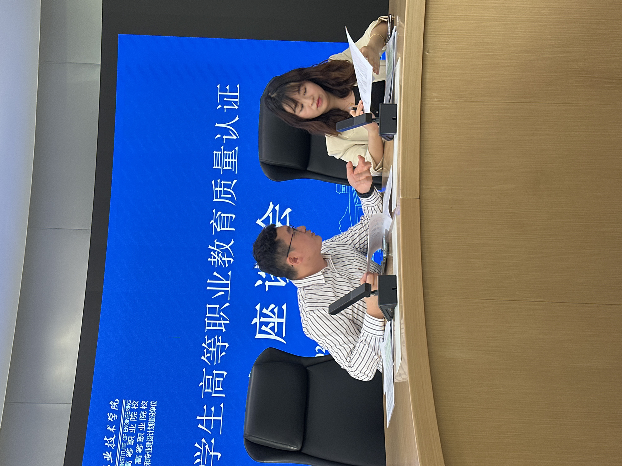 The image depicts an indoor meeting or symposium taking place in Jiangjin, China, with two individuals seated at a light-colored wooden table. A man, wearing a striped shirt and glasses, is positioned on the left side of the table, actively gesturing with his right hand while looking at papers. A microphone is placed in front of him. Opposite him, on the right, a woman with long, dark hair, dressed in a light-colored jacket, is reviewing documents with a focused expression.
Behind them, a large blue screen serves as a backdrop, displaying white text in both Chinese and English. The prominent Chinese text reads: "学生高等职业教育质量认证" (Student Higher Vocational Education Quality Certification) and "座会" (Symposium/Meeting), with the number "12" also visible. Additional Chinese text includes "二业技术学院" (potentially part of an institute's name like "Institute of Technology"), "高等职业院校" (Higher Vocational College/Institute, appearing twice), and "三和专业建设计划建设单位" (Sanhe Professional Construction Planning and Construction Unit). The English text "INSTITUTE OF ENGINEERING" is clearly visible, along with parts of other English text like "HIGHER VOCATIONAL COLLEGE" which corresponds to the Chinese.
The scene suggests a formal discussion or review session, likely related to quality certification for higher vocational education, given the visible text. Both participants appear engaged in the process. The lighting is bright and even, characteristic of an indoor conference setting.