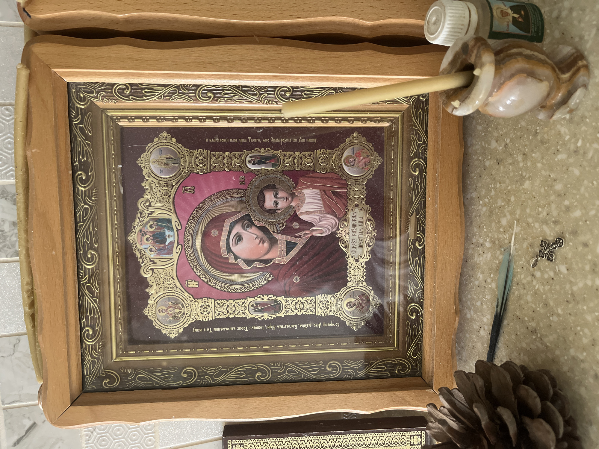 The image shows a framed icon of the Virgin Mary and Child, likely an Orthodox Christian icon, resting on a speckled countertop. The icon is adorned with gold-colored ornamentation and is behind glass. To the left of the icon, a large pinecone is visible, and next to it is a single blue and green feather and a small silver cross pendant. To the right of the icon, a lit candle stands in a small, marbled candle holder. Behind the candle holder is a small bottle with a white cap, featuring a label with a picture of a person in religious attire. The background consists of a tiled wall and wooden paneling. The scene is indoors, likely on a shelf or table, in a domestic setting. The specific location in the city of Kommunarka, Russia, is not discernible from the image itself.