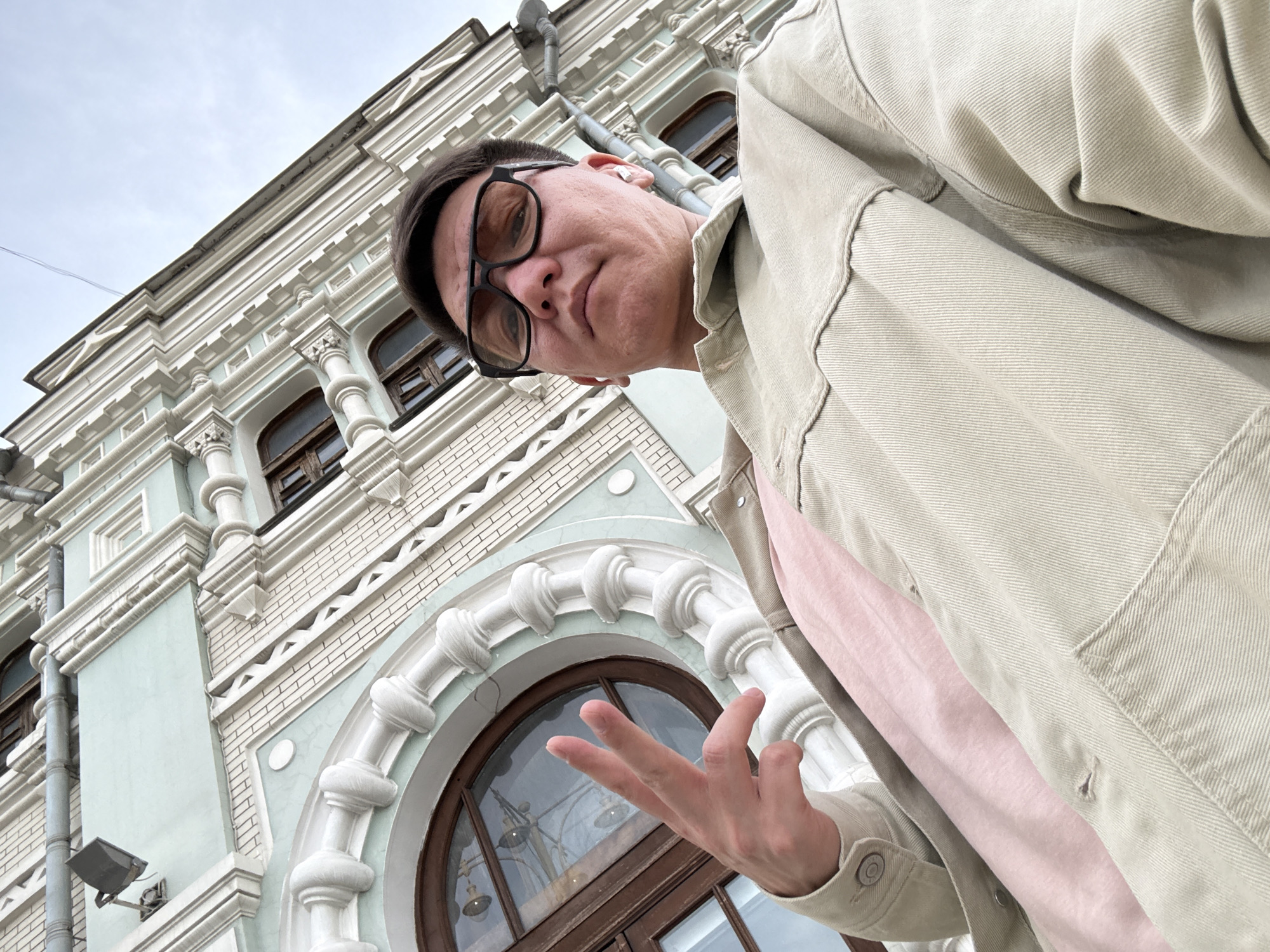 A low-angle shot features a person with short dark hair, wearing black-rimmed glasses and a light-colored jacket over a pink shirt. The individual is making a hand gesture. Behind them is a building with ornate architectural details, including arched windows and decorative moldings. The building's color scheme is a combination of light blue-green and white. The sky is overcast. The location is Moscow, Russia, as evidenced by the distinctive architectural style of the building.