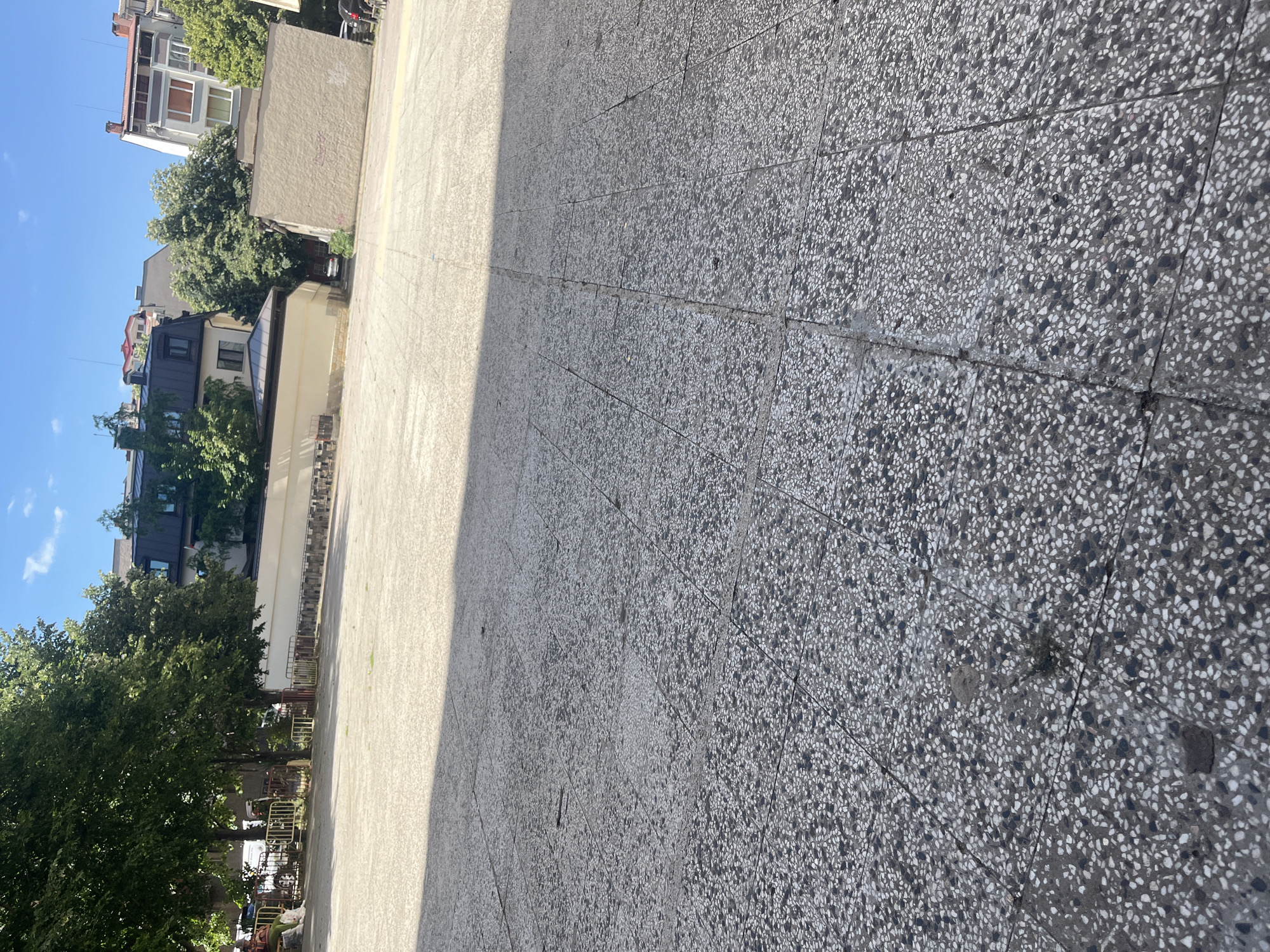 The image shows a sunlit, open paved area in Stara Zagora, Bulgaria. The ground is composed of large, square-shaped concrete tiles with a speckled texture. A distinct shadow diagonally bisects the lower portion of the frame, indicating a strong light source from the upper left. In the background, trees with lush green foliage are visible, along with buildings of varying architectural styles. Some buildings appear to be residential, with visible windows and rooflines. A low, light-colored wall with a fence in front of it stretches across the mid-ground. The sky is a clear blue with a few wispy white clouds. The scene appears to be an outdoor public space, possibly a courtyard or plaza. No individuals are clearly depicted in the image, though it is possible there are people obscured by the distance or foliage.