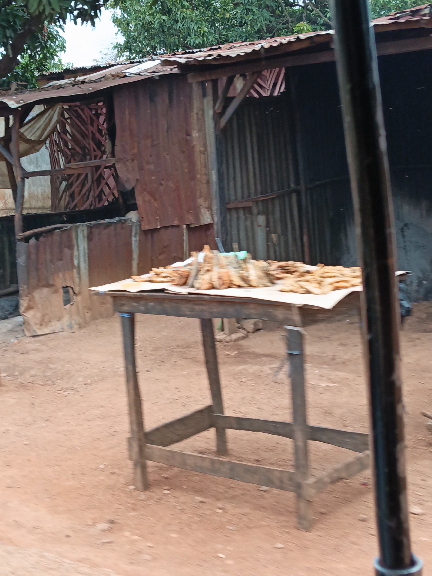 A rustic wooden table is laden with a large display of dried fish, piled high on a white sheet of paper. The table itself is constructed from rough-hewn wood, with a simple, utilitarian design. In the background, a ramshackle structure made of corrugated metal and weathered wood stands, its roof patched and uneven. Lush green foliage peeks over the top of the structure, suggesting a humid, possibly tropical environment. The ground is a dusty, reddish-brown earth. A dark, vertical pole, possibly part of a fence or a building's support, is prominently positioned in the foreground, slightly obscuring the view of the scene. The overall impression is one of a simple outdoor market stall in a less-developed area.