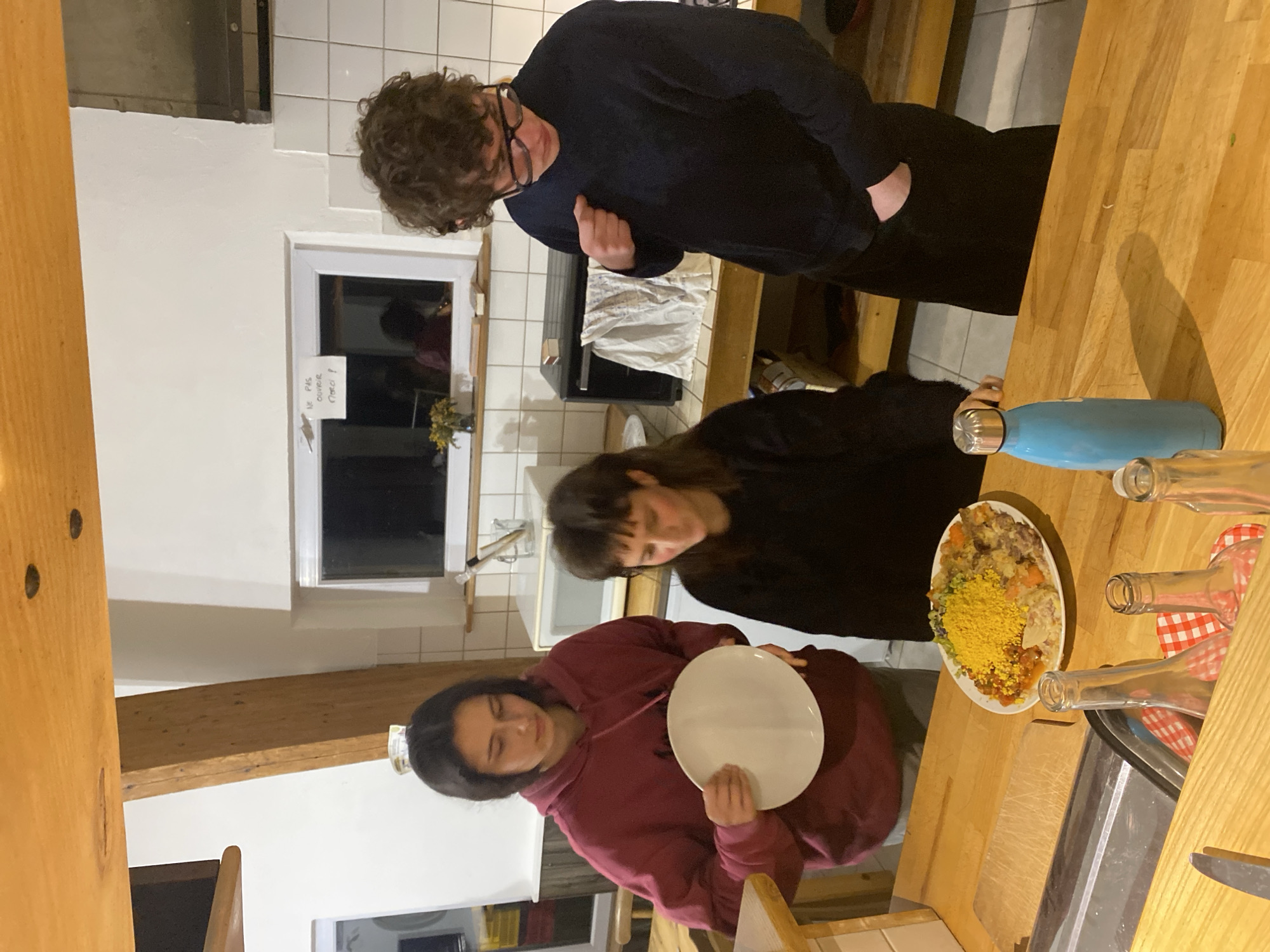 Three people are gathered around a wooden countertop in a kitchen. One person, wearing a dark blue sweater and glasses, stands to the right of a window with a sign taped on it. Another person, in a black sweater, is near a plate of food. A third person, wearing a burgundy hoodie, holds a white plate. There are several empty glass bottles and a blue water bottle on the countertop. The location is the city of Cornillon-en-Trièves, France.