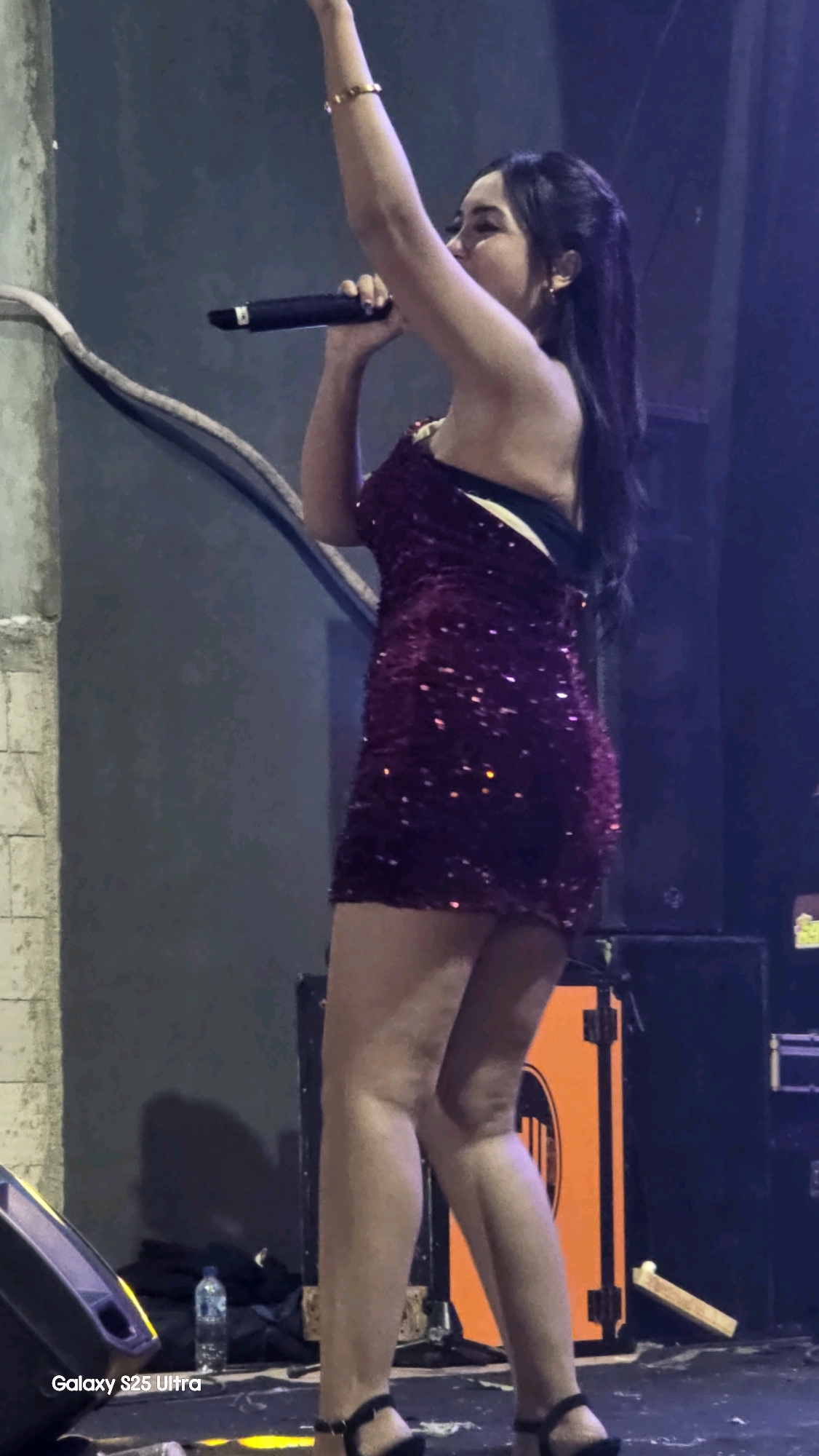 A woman wearing a sequined dress and high heels is singing into a microphone on a stage in Jaken, Indonesia. She is holding the microphone with her right hand, and her left arm is raised in the air. The stage is lit with bright lights, and there are speakers and other equipment in the background. The woman has long, dark hair and is wearing makeup. The image is taken from a low angle, and the woman is in focus. The background is blurred, and the lighting is dim.
