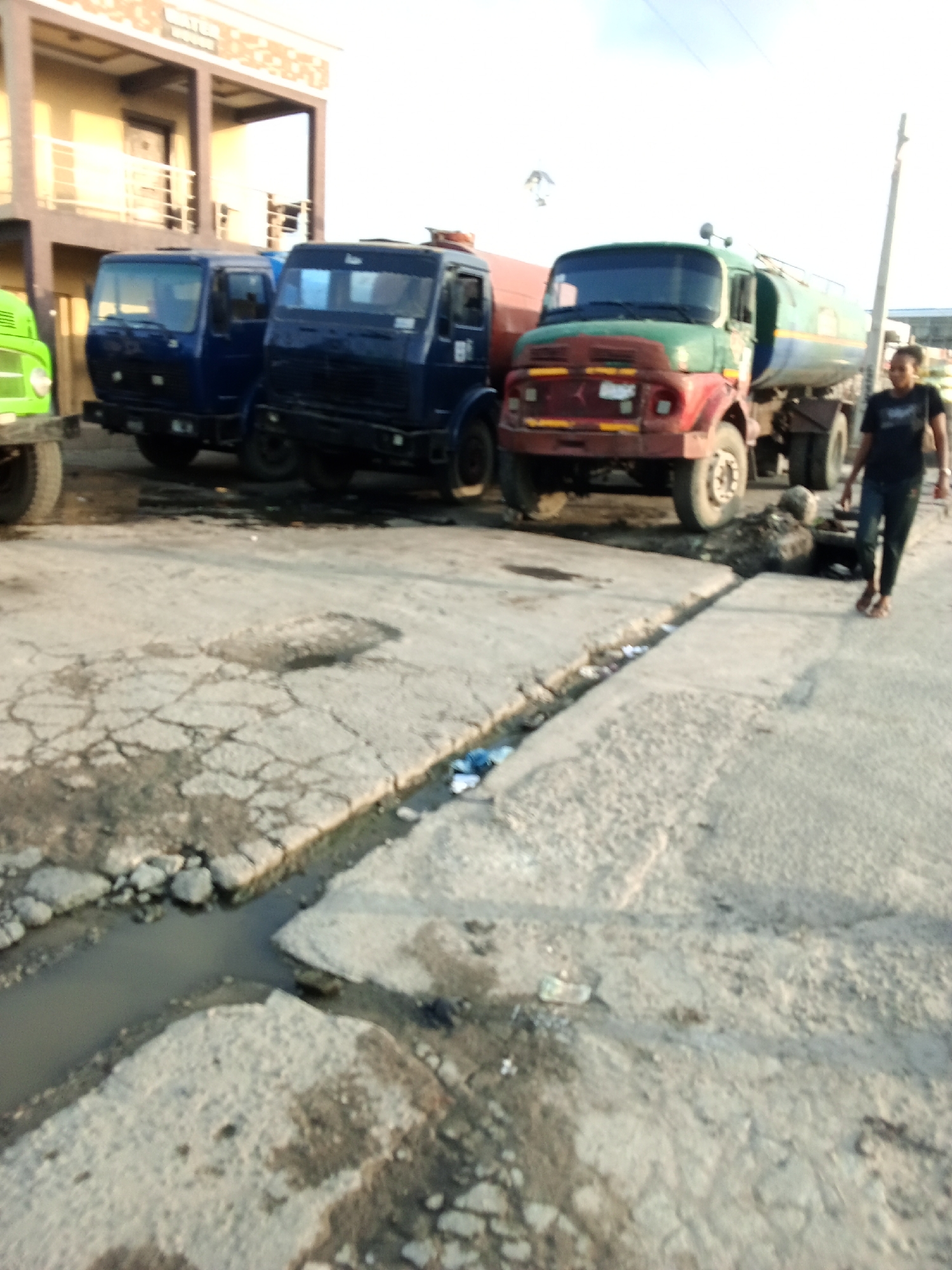 The image depicts a street scene in Ado, Nigeria. Three trucks are parked near a two-story building. The trucks are green, blue, and red. A person is walking in the street. There is a drainage ditch along the side of the street, partially filled with water and some litter. The pavement shows signs of wear and damage.