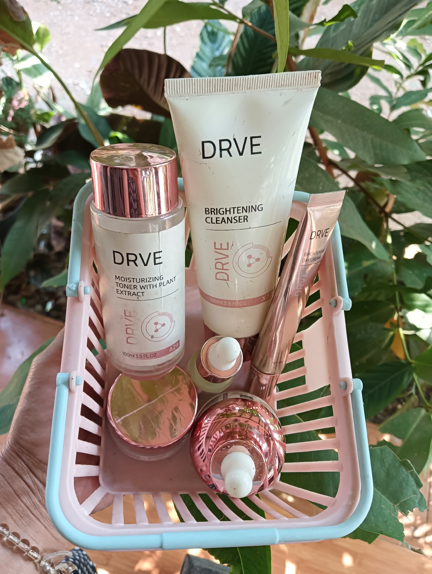 In Cluwak, Indonesia, a hand holds a small pink and blue plastic basket filled with various DRVE skincare products. The basket contains a large tube of DRVE Brightening Cleanser, a clear bottle of DRVE Moisturizing Toner with Plant Extract (100ml/3.5 FL.OZ), a rose gold tube of DRVE Brightening Day Cream, a small dropper bottle with a white cap and a rose gold accent, and a larger rose gold dropper bottle with a white top. The products are arranged with the cleanser and toner bottle in the back, the day cream tube and smaller dropper bottle in the middle, and the larger dropper bottle in the front. The basket and its contents are positioned in front of lush green foliage.